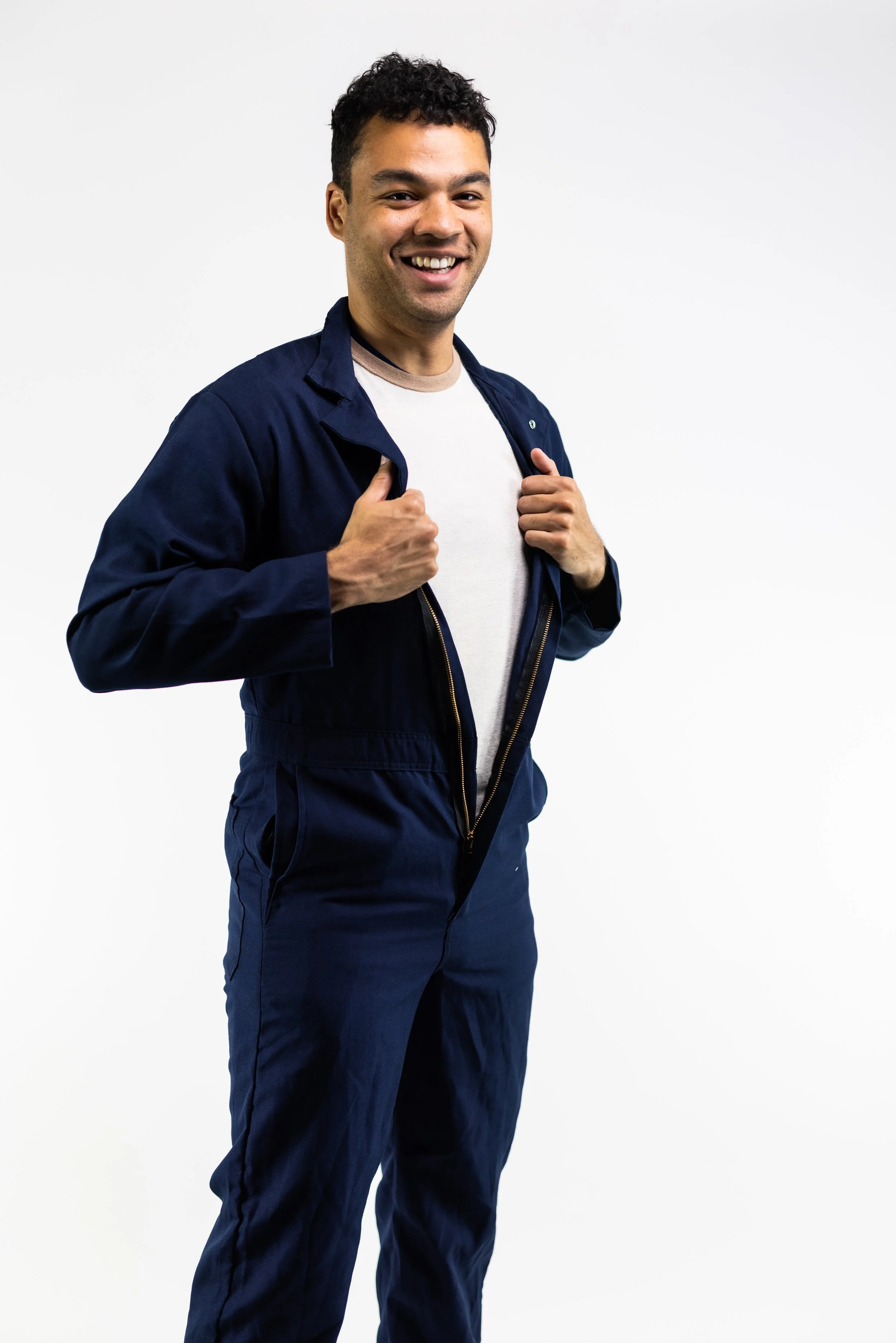 A young man with dark curly hair smiling and adjusting his navy blue jumpsuit, against a plain white background.