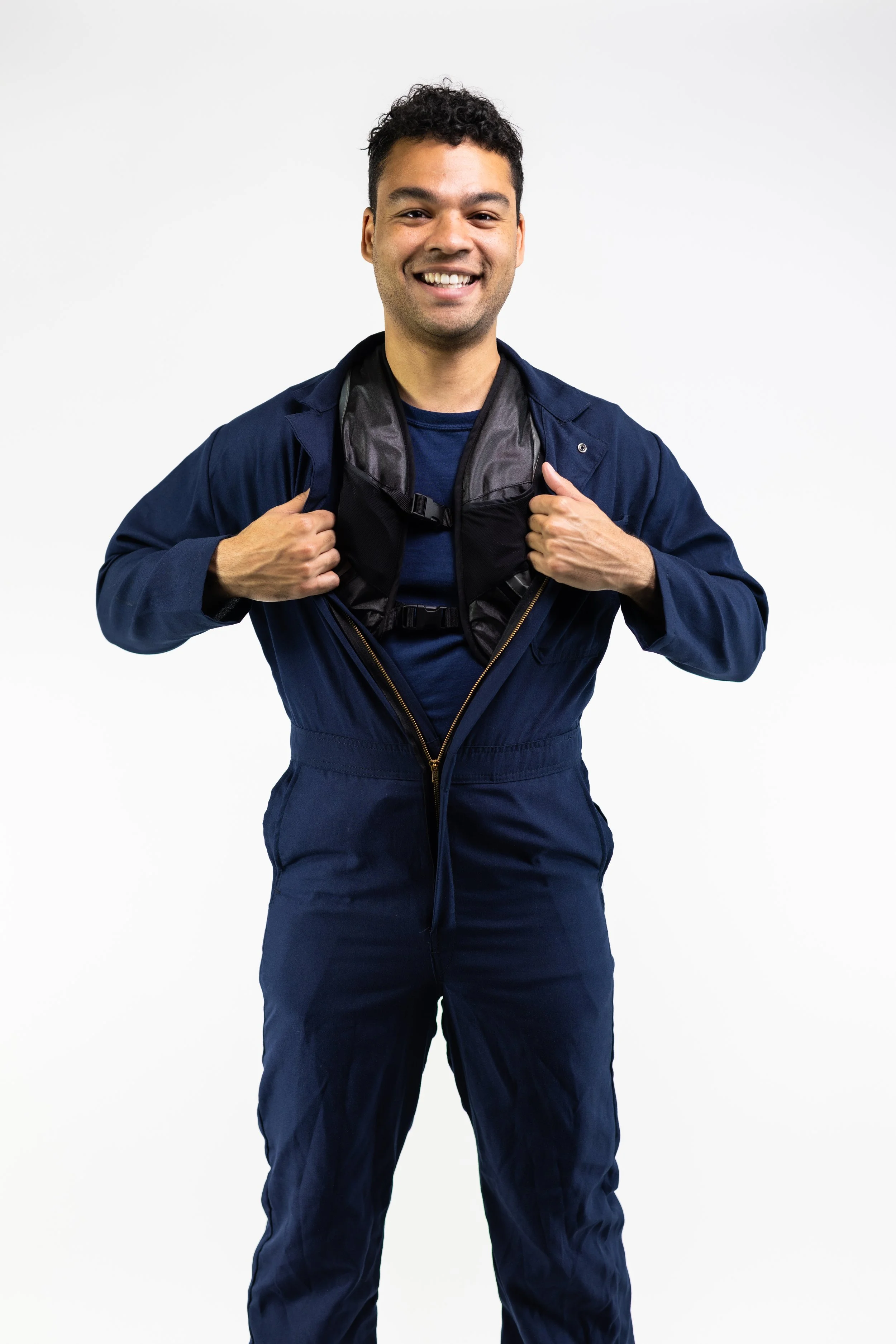 A smiling man in a navy jumpsuit holding open the collar of his jumpsuit to reveal a black vest underneath, standing against a plain white background.