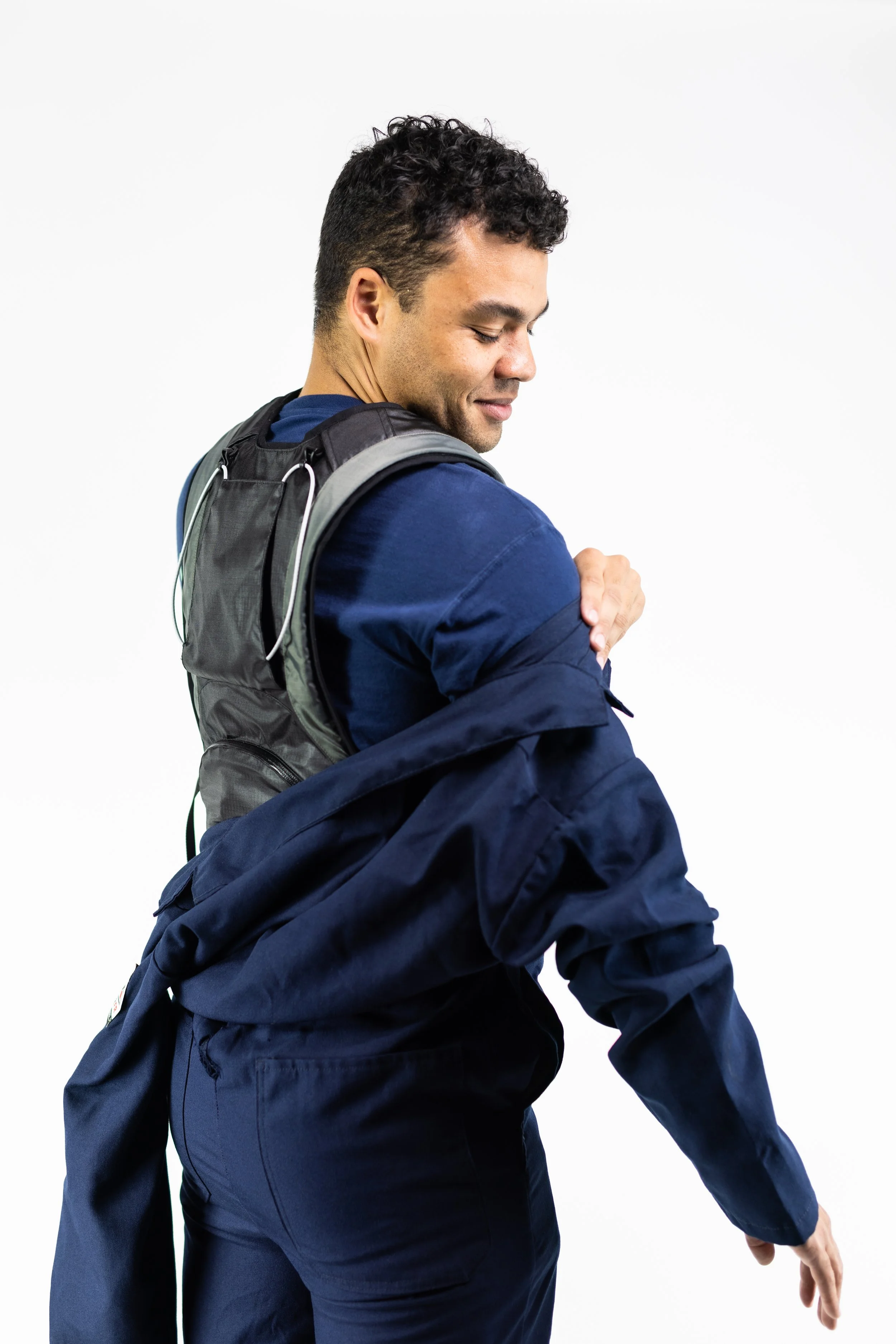 A man wearing a blue jacket and backpack smiling and looking down while adjusting his sleeve against a plain white background.