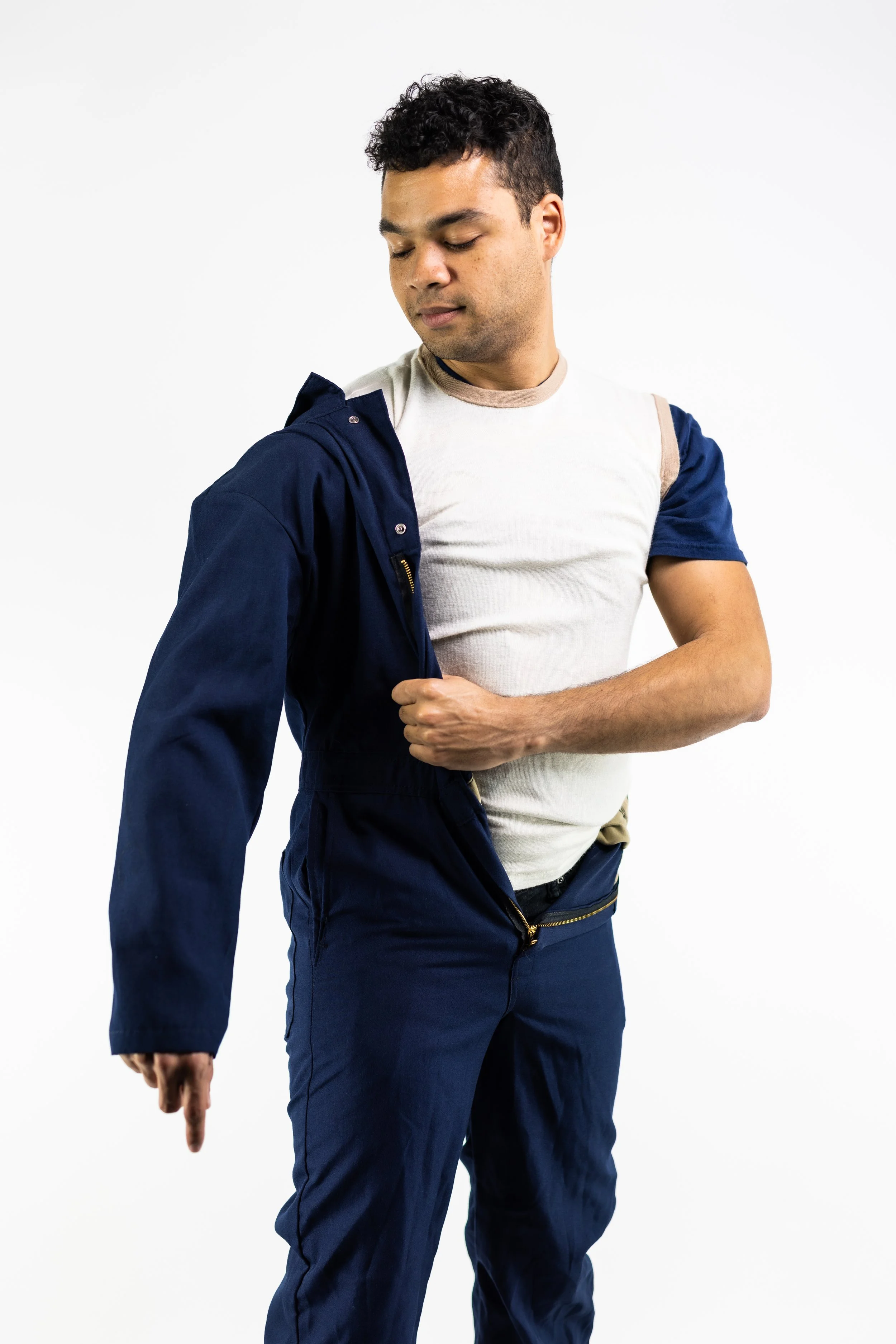 A young man with short curly hair is wearing a white t-shirt, a navy blue jacket, and matching navy blue pants. He is looking down and pointing at his leg with his left hand, standing against a plain white background.