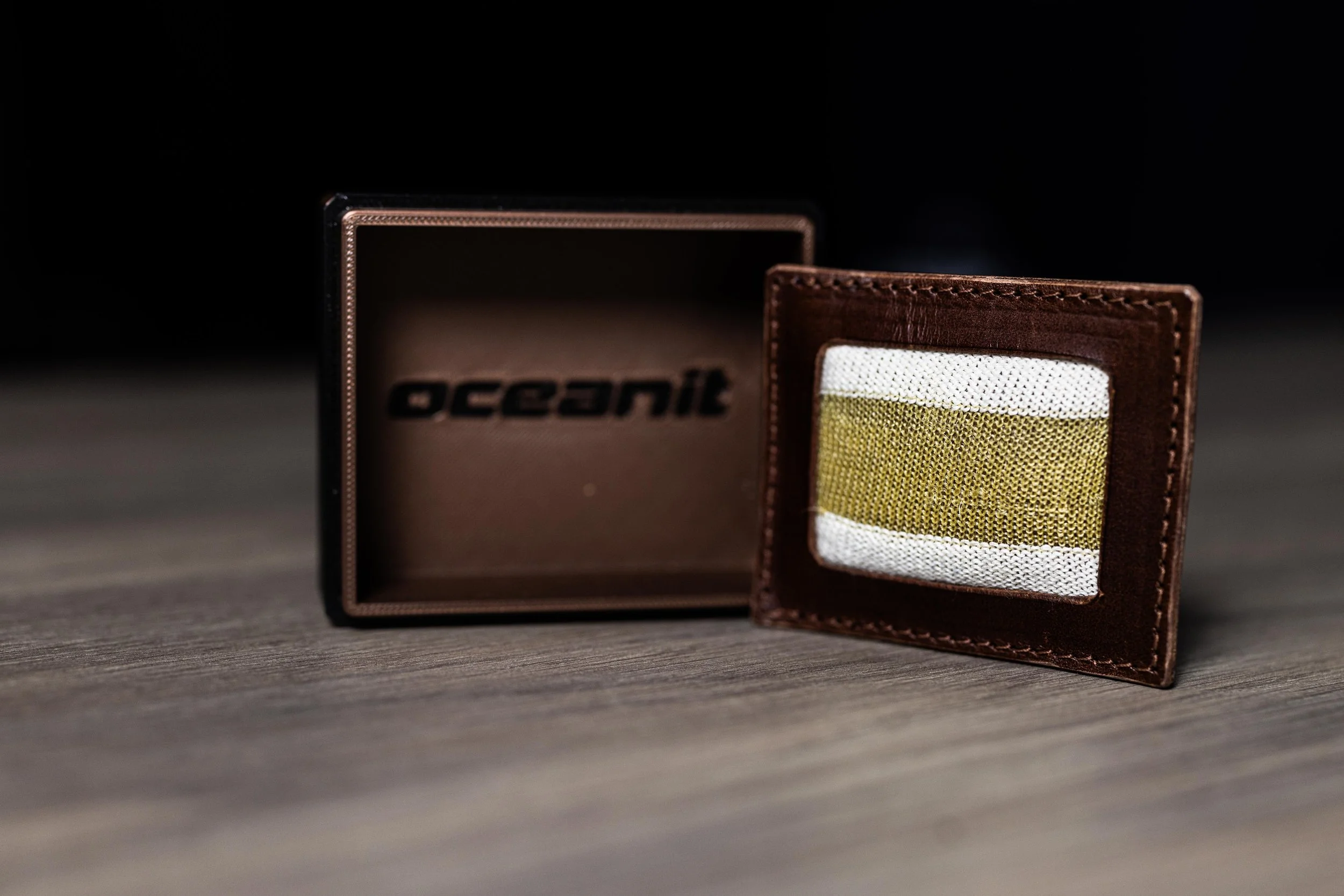 A brown leather wallet with a windowed ID slot and a white and yellow fabric patch inside, placed in front of a black and brown box with the word 'oceanic' on it, on a wooden surface.