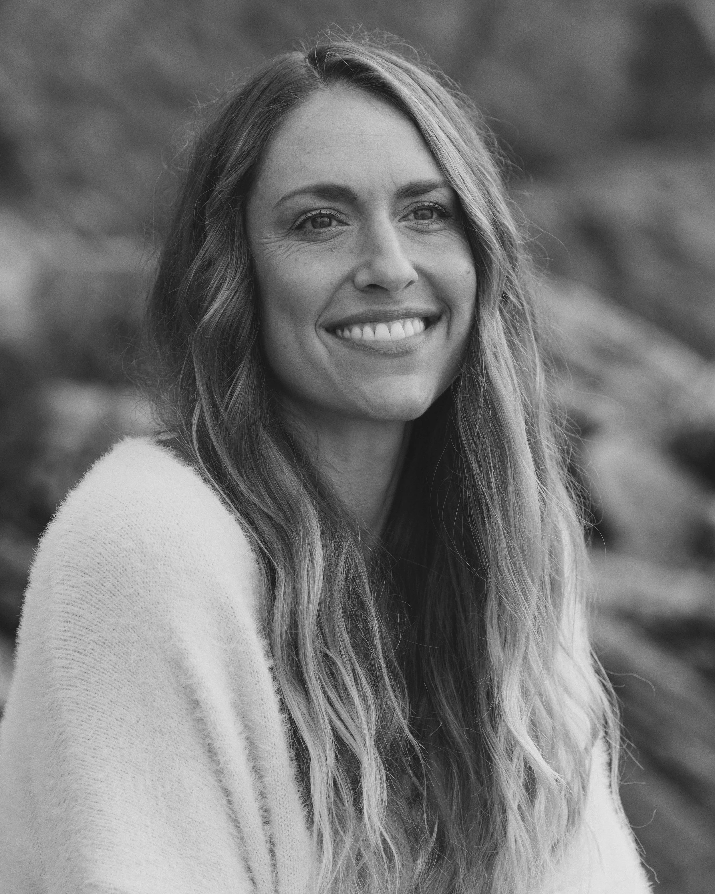 A black-and-white portrait of a woman with long hair, smiling, outdoors with blurred nature background.