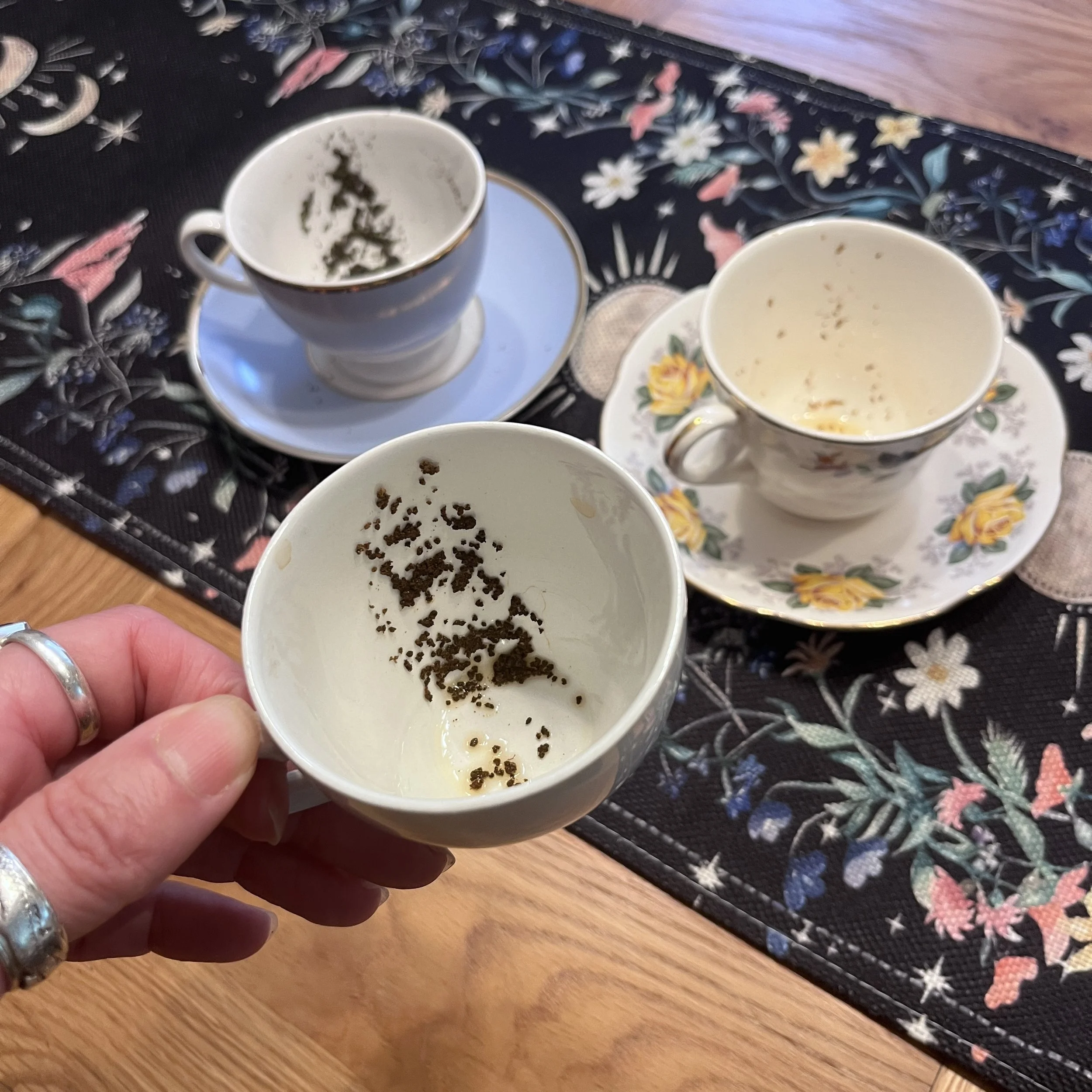 A person holding a cup with tea leaves inside, with other cups and flower-patterned saucers on a table. The table has a floral tablecloth with a colorful floral design.