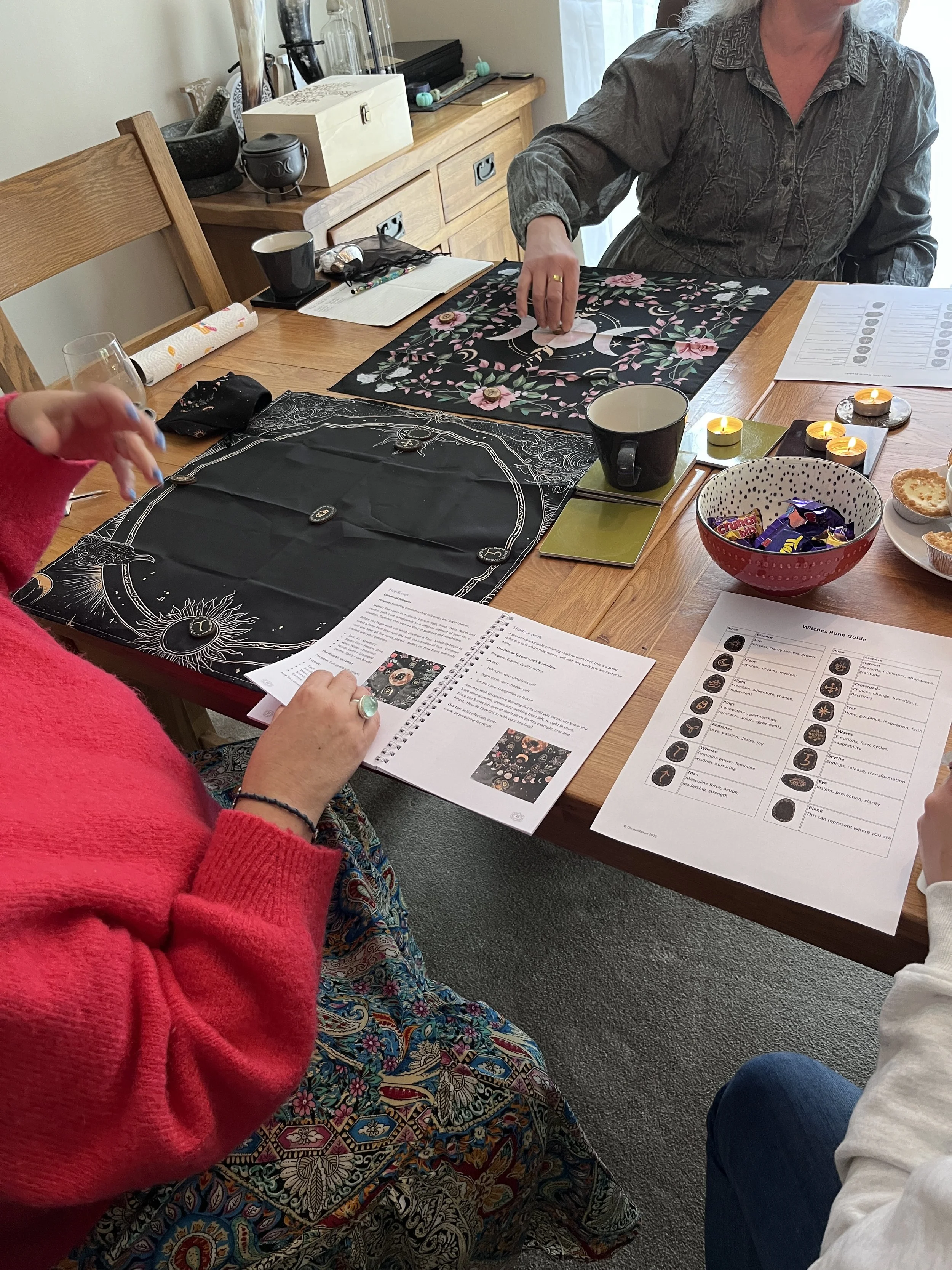 People seated around a wooden table engaged in a discussion or activity with Tarot or oracle cards, candles, snacks, and printed guides on the table.