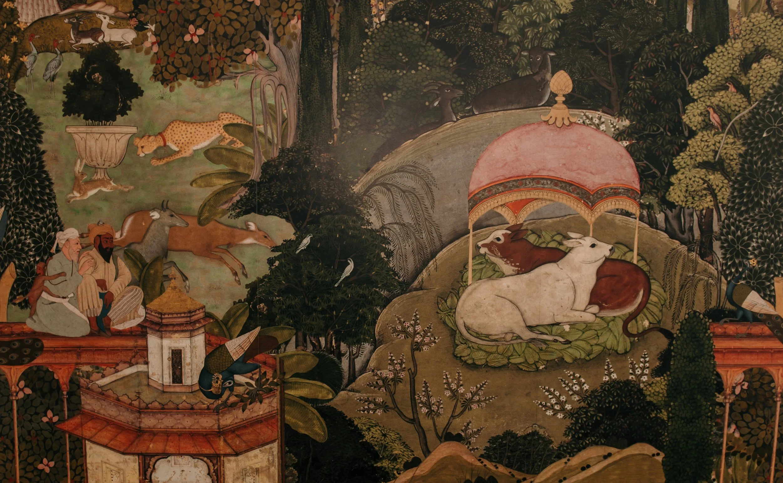 A detailed, colorful mural depicting a variety of animals including cows, deer, rabbits, birds, and a leopard, with two human figures, trees, and a fountain with a bird, set in a lush, wooded landscape.