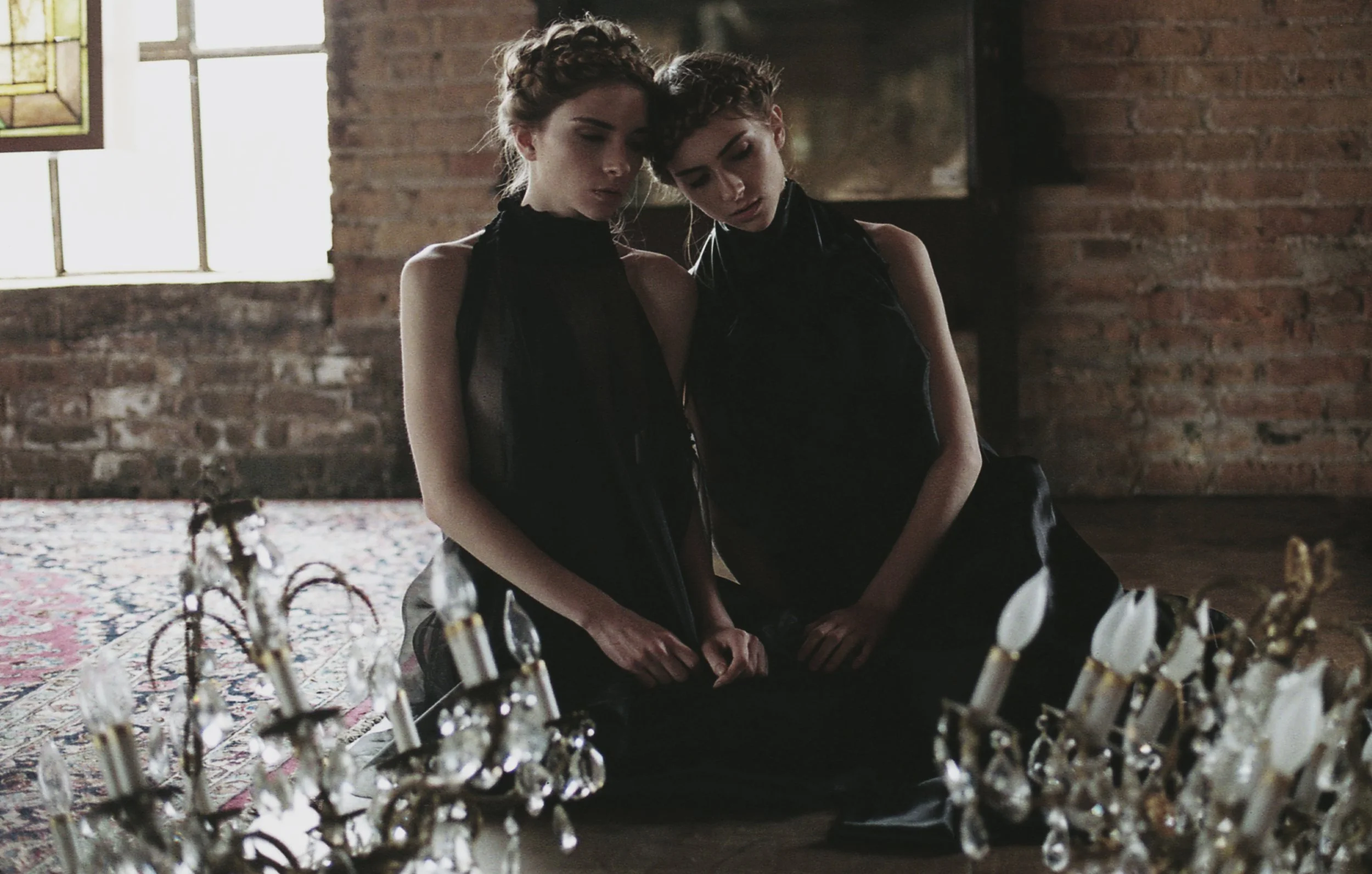 Two young women with short, curly hair, dressed in black, sit close together on the floor, looking down in a dimly lit room with an exposed brick wall and a stained glass window. A large ornate chandelier with white candles and hanging crystals is in