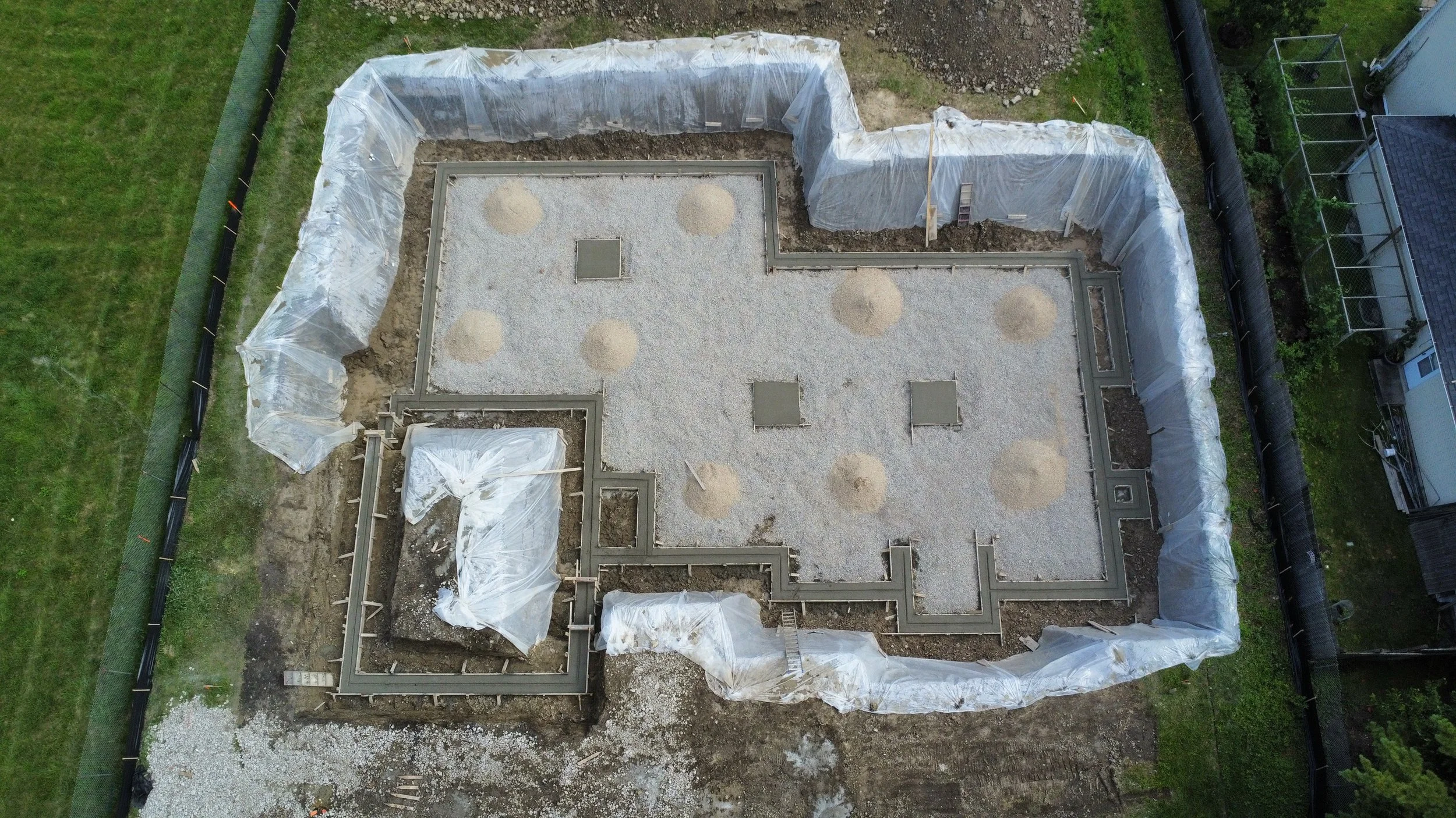 Coorlas Architecture aerial view of a building foundation under construction, with gravel and concrete forms, surrounded by plastic sheeting and green yard areas.
