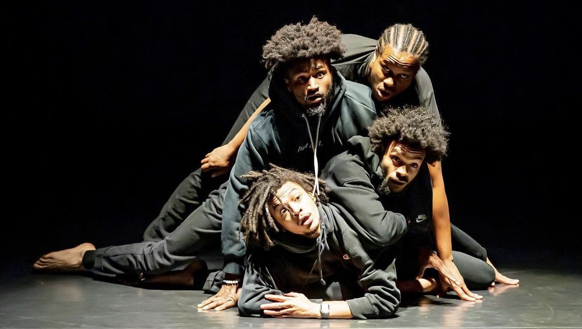 Four people in black casual clothing performing a dance or theatrical pose on a dark stage, intertwined and on the floor.