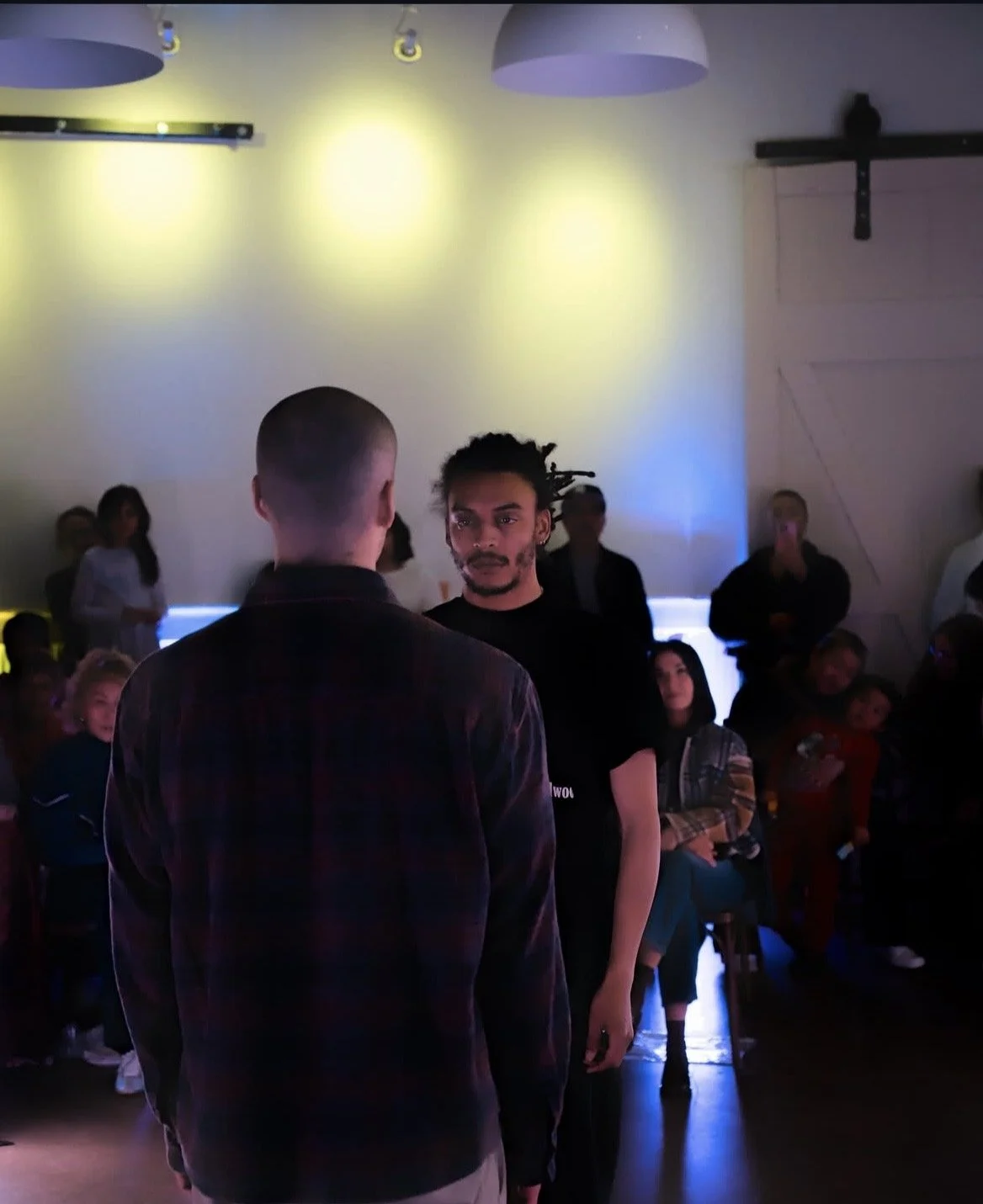 Two men face each other in the foreground of a dimly lit room with an audience watching. The man facing away has a buzzed haircut, and the man facing forward has dreadlocks and facial hair.