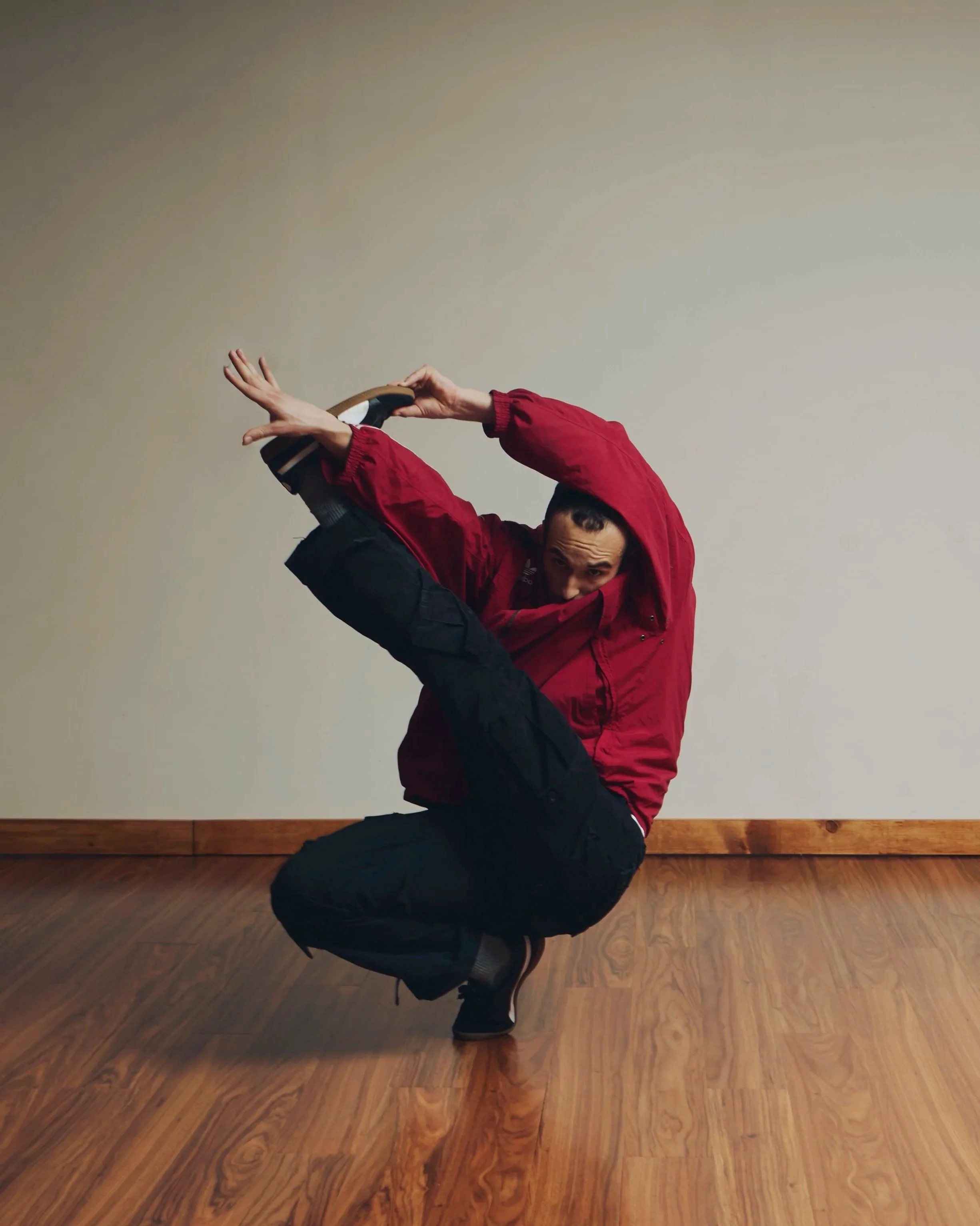 Person in red jacket performing a breakdancing move indoors on wooden floor, with plain wall background.
