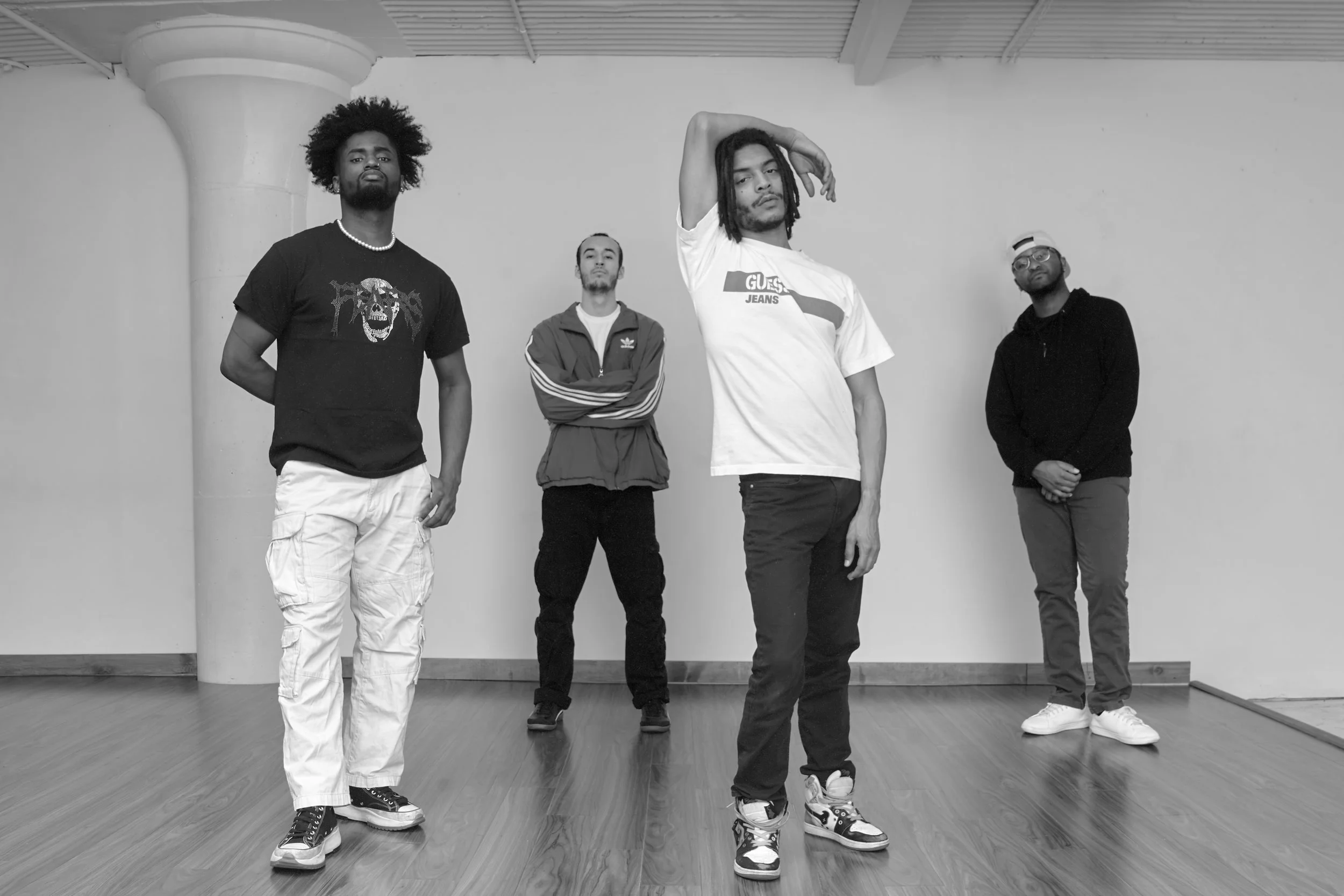 Four men standing on a wooden floor against a plain wall, posing for a photo. One man in front with a hand on his head, wearing a branded T-shirt and sneakers. Behind him, another with arms crossed, wearing a jacket and Adidas track pants. To the left, a man with curly hair and a T-shirt, and on the far right, a man in a hoodie with glasses, all looking towards the camera.
