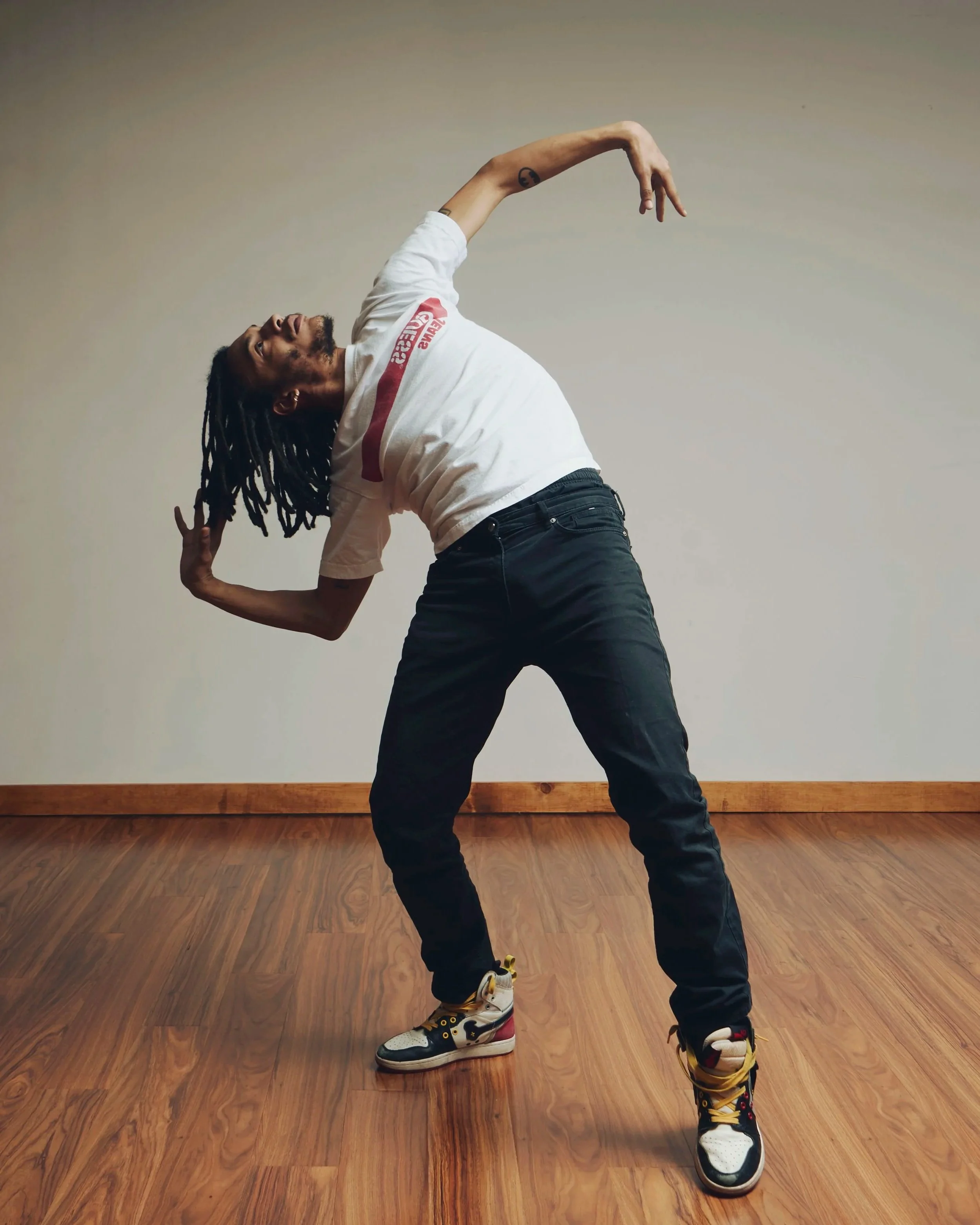 A man with dreadlocks in a dance pose, leaning to the side with one hand behind his head and the other arm extended, standing on a wooden floor against a plain wall.