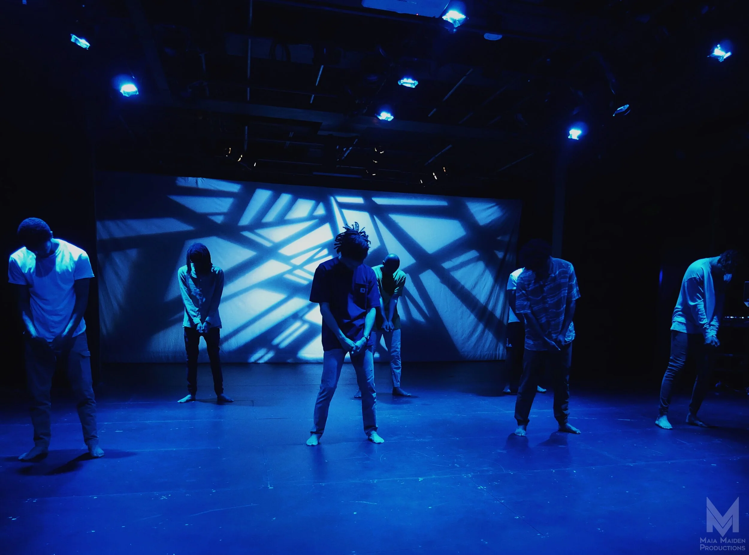 Six dancers are practicing on a dark stage illuminated by blue lights, with shadows cast on a large backdrop behind them.