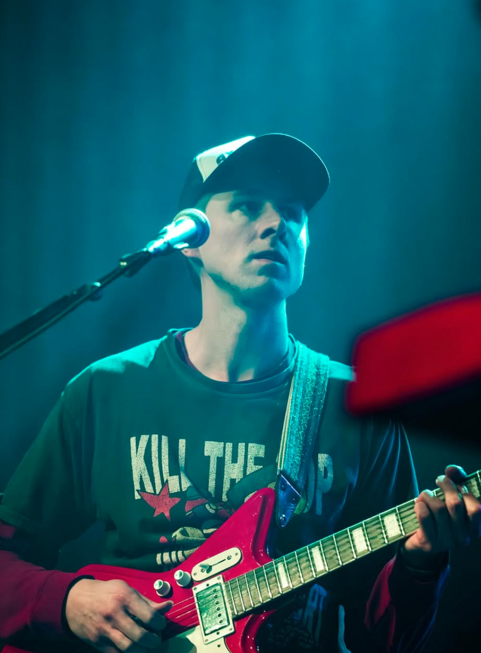 A young man wearing a black cap and a black T-shirt with a graphic and text, playing a red electric guitar in a dimly lit setting, with a microphone in front of him.