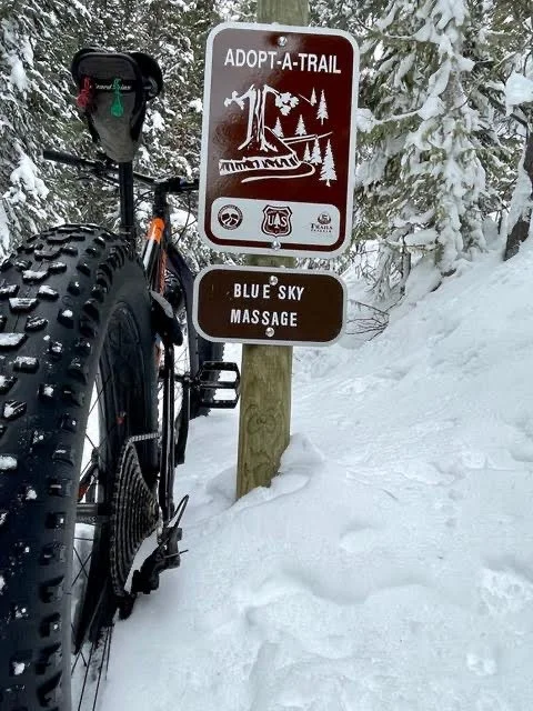 A mountain biking trail sign with Blue Sly Massage written on a snowy forest. The top sign reads "Adopt-a-Trail" with a graphic of a mountain, trees, and a bike. The bottom sign says "Blue Sky Massage." A mountain bike is parked beside the signs.