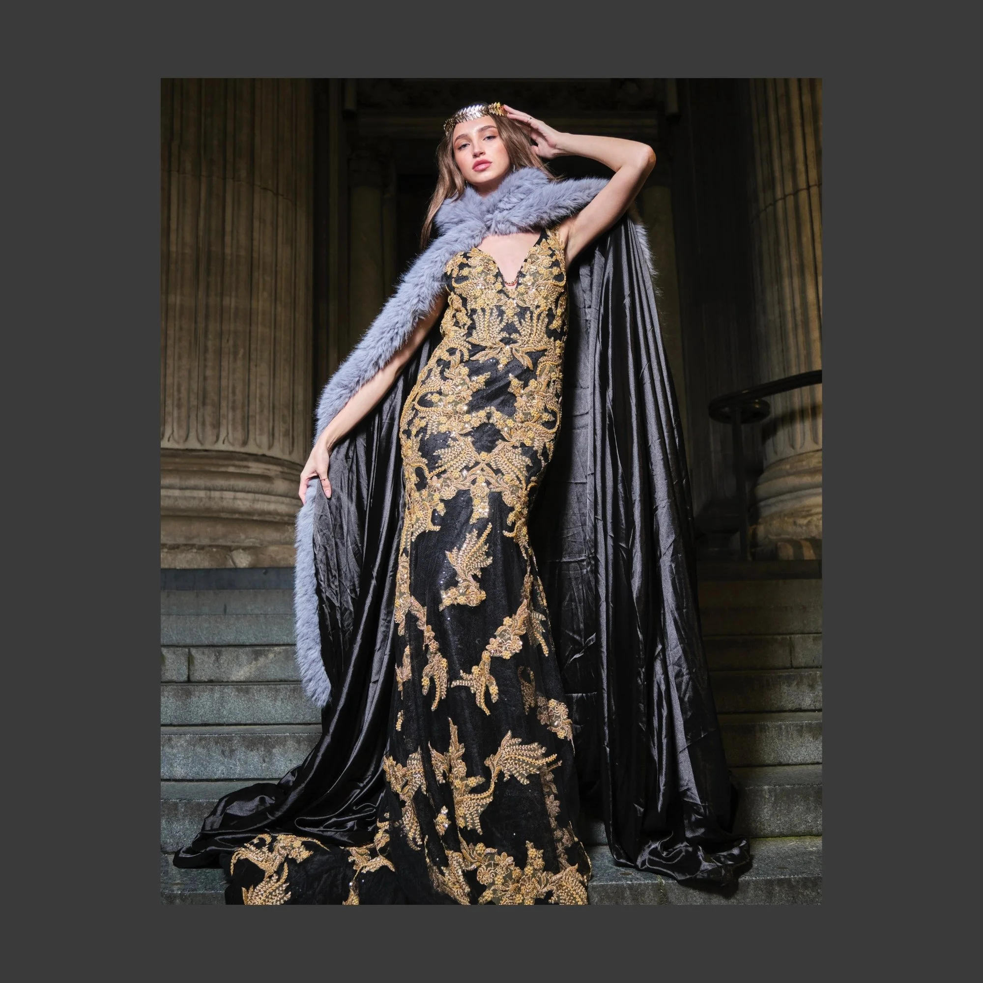 A woman in a luxurious gold and black embroidered gown with a plunging neckline, draped in a black satin cape and a gray fur stole, standing on stone steps in front of grand columns, wearing a decorative headband.