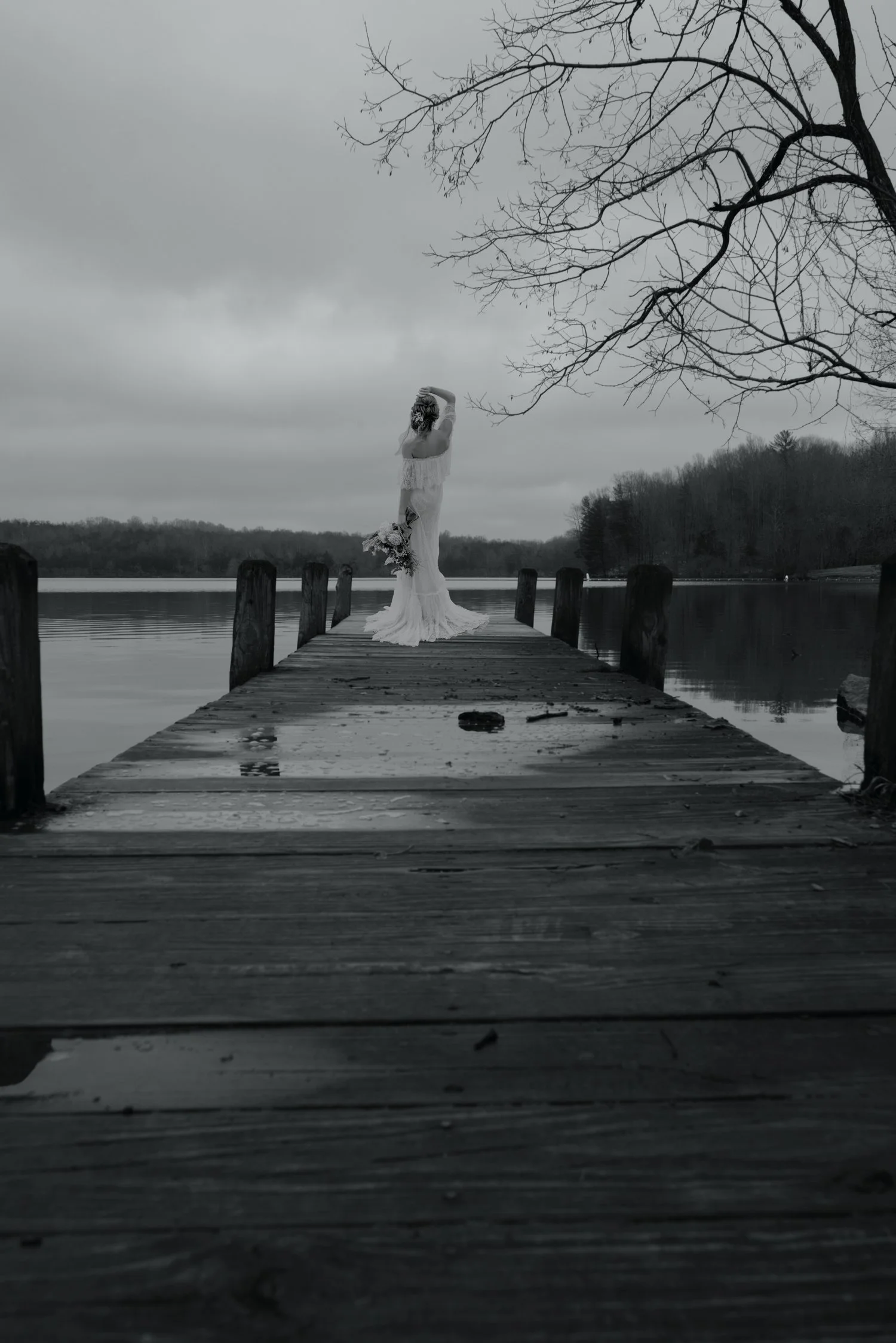 Black and white photo of a woman in a wedding dress holding a bouquet, standing at the end of a wooden dock on a calm lake with a cloudy sky, leafless tree branches overhead, and distant forest in the background.