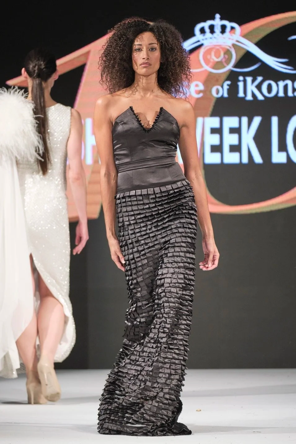 Model walking on runway during a fashion show, wearing a black satin sleeveless top with a ruffled hem and matching black textured wide-leg pants.
