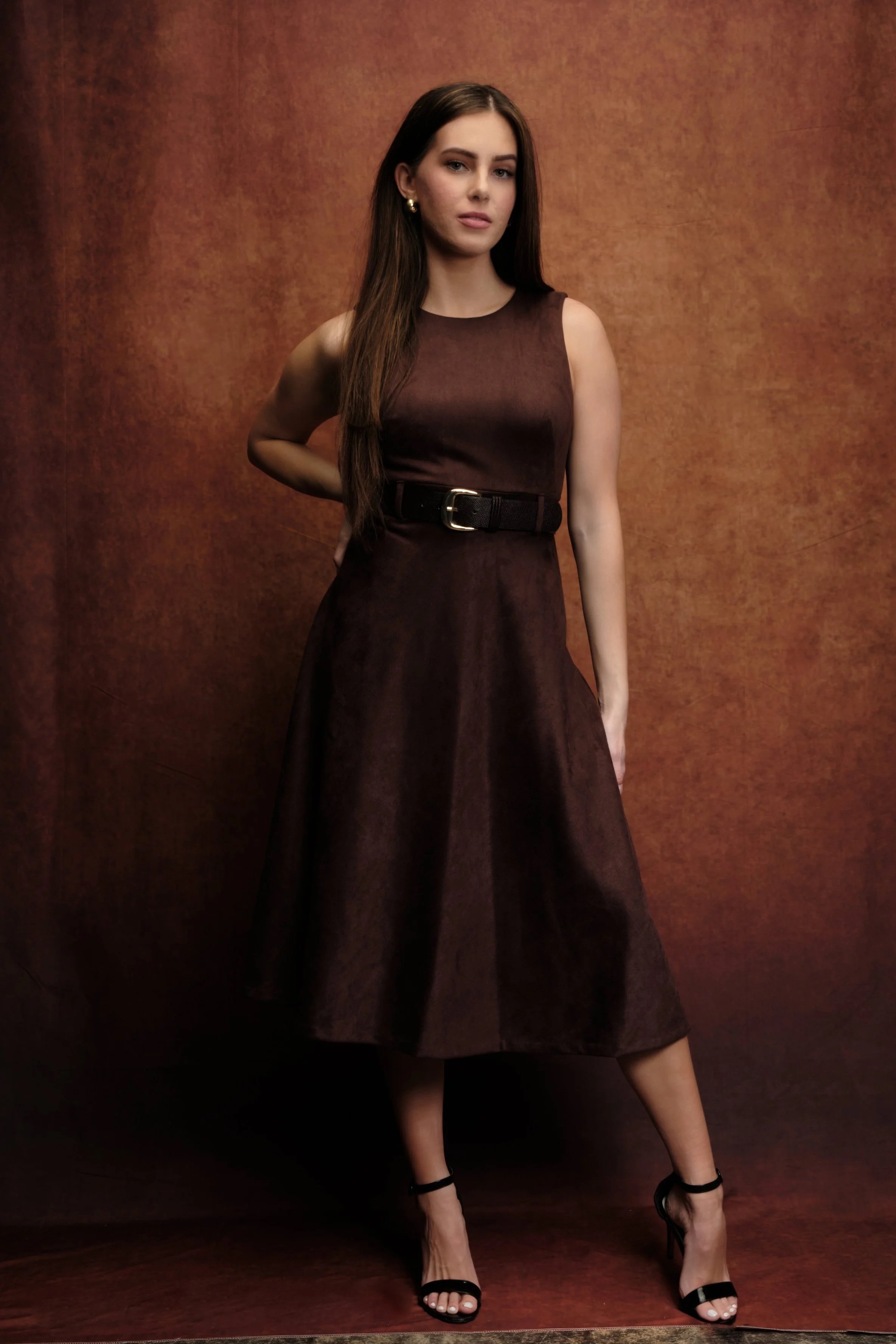 A young woman with long brown hair wearing a sleeveless dark brown midi dress with a black belt and black high-heeled sandals, standing against a textured brown background.