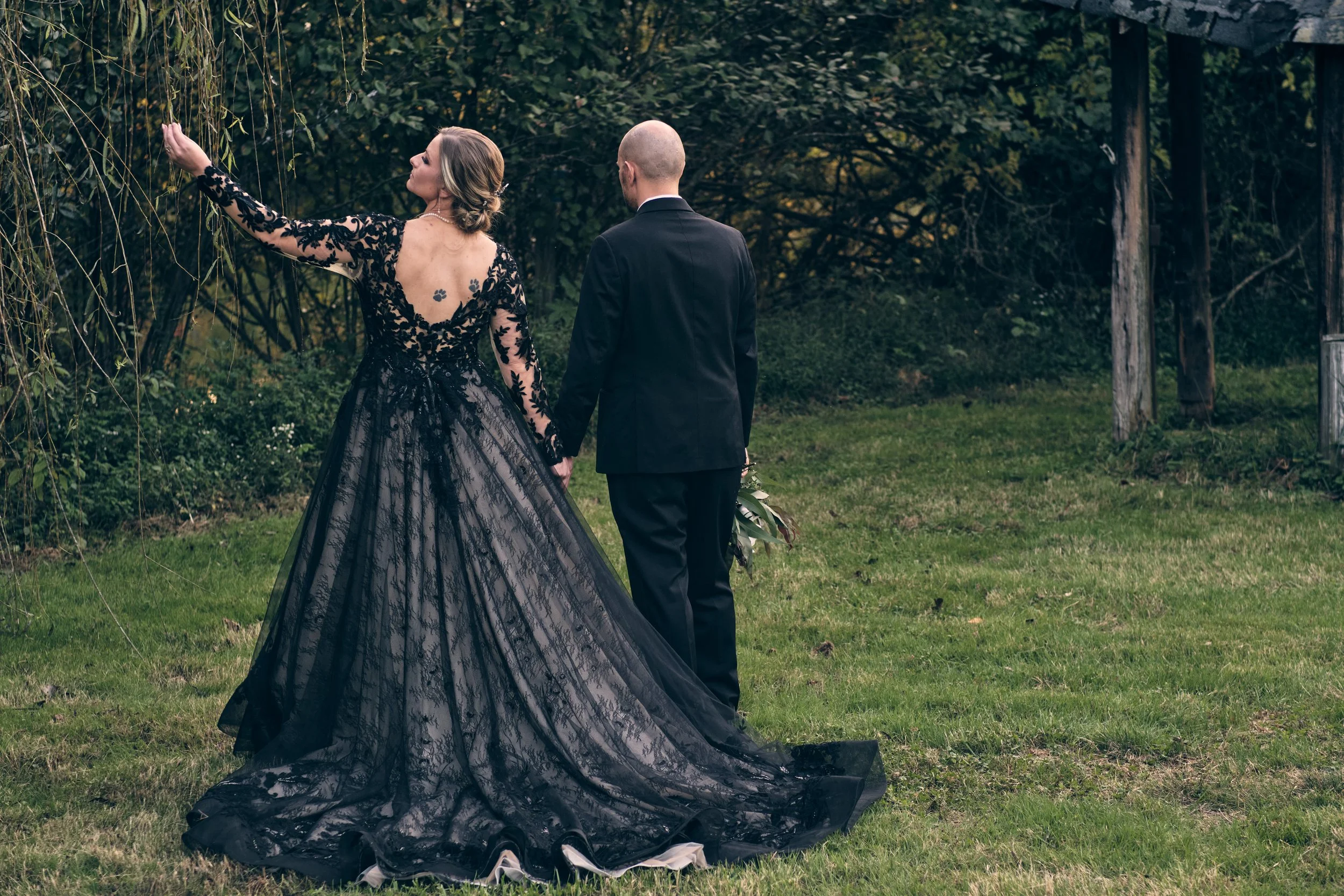 A woman in a black lace gown with a long train and a man in a black suit holding hands outdoors near trees, with the woman reaching out and touching tree branches and the man holding a bouquet of flowers behind her.