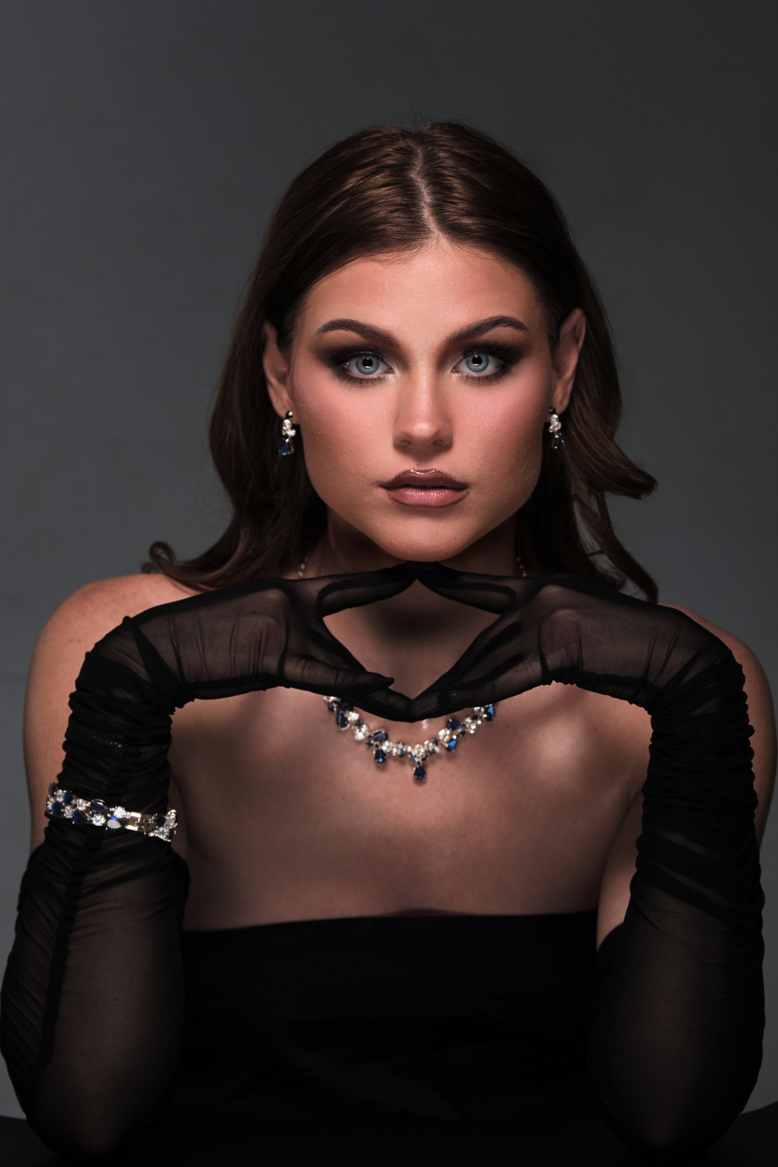 A woman with blue eyes and dark brown hair styled in loose waves wearing black gloves, elegant jewelry including a necklace and earrings, and a strapless black dress, posing with her hands near her face against a dark background.