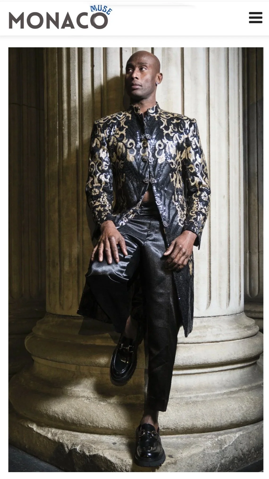 A man wearing a black and gold patterned blazer, black pants, and black shoes, sitting on a stone step in front of a large column.