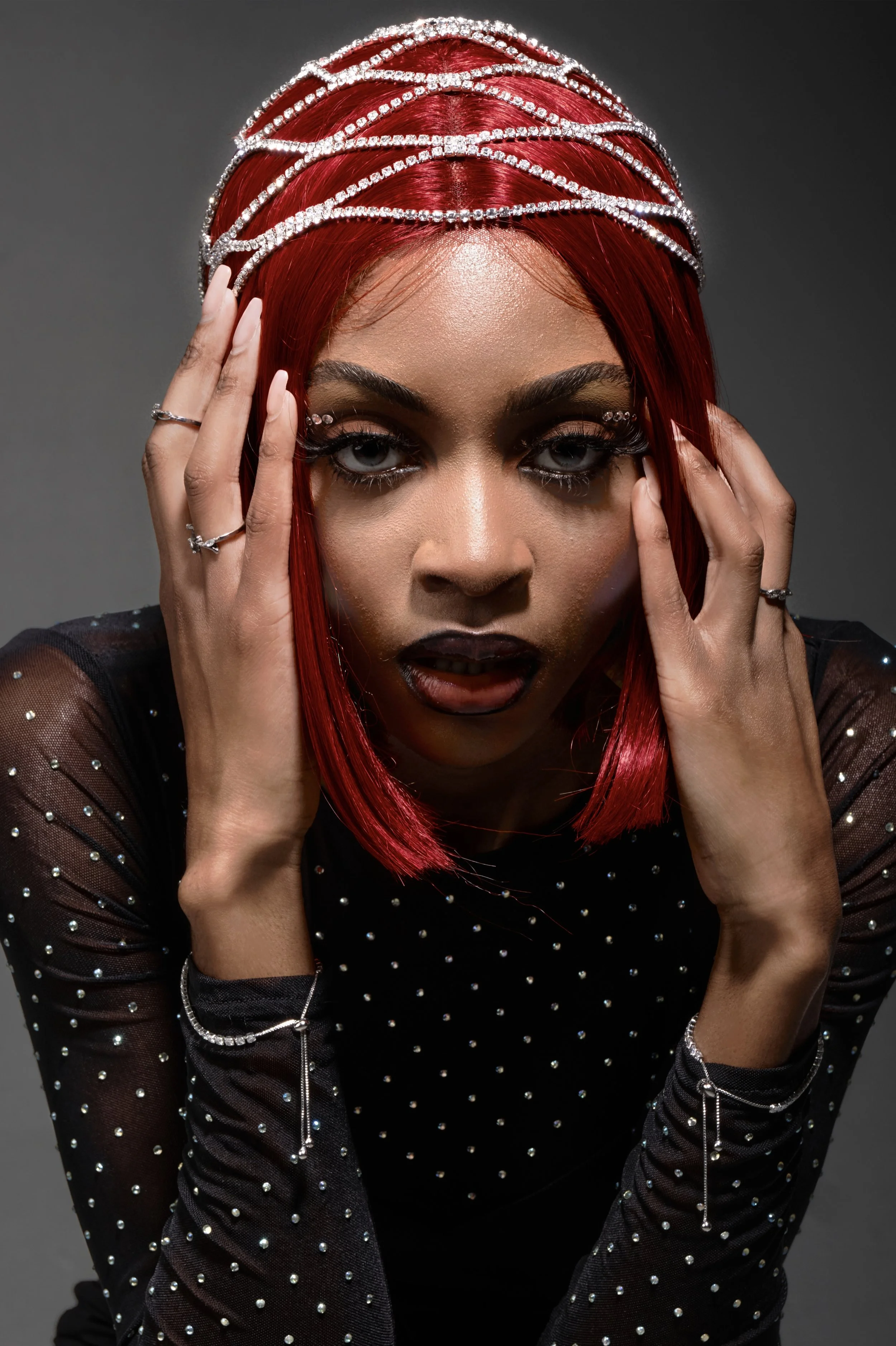 A woman with red hair and silver jewelry, wearing a black, glittery top, posing with her hands around her face against a gray background.