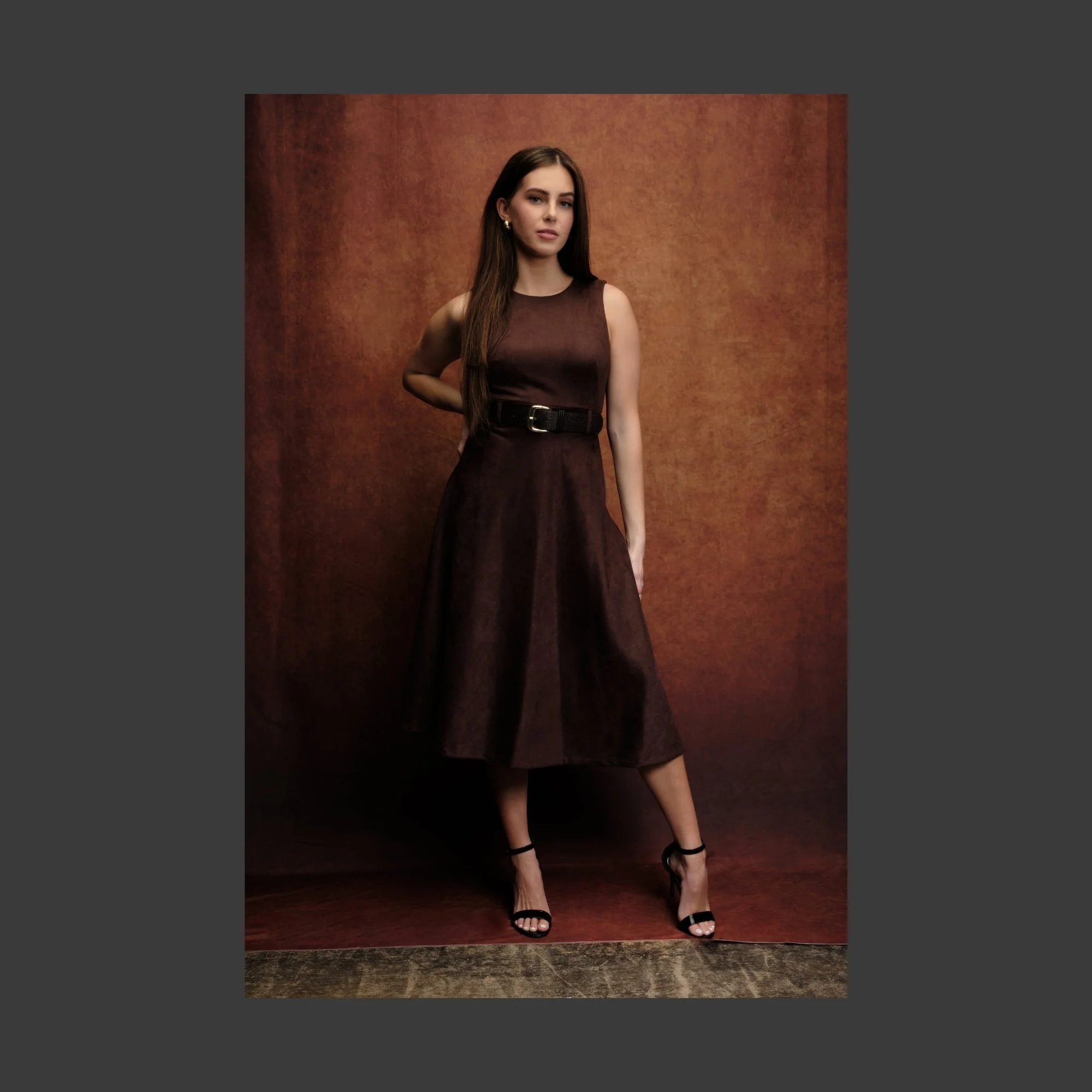 A young woman with long brown hair wearing a sleeveless dark brown dress with a black belt and black high-heeled sandals standing against a brown background.