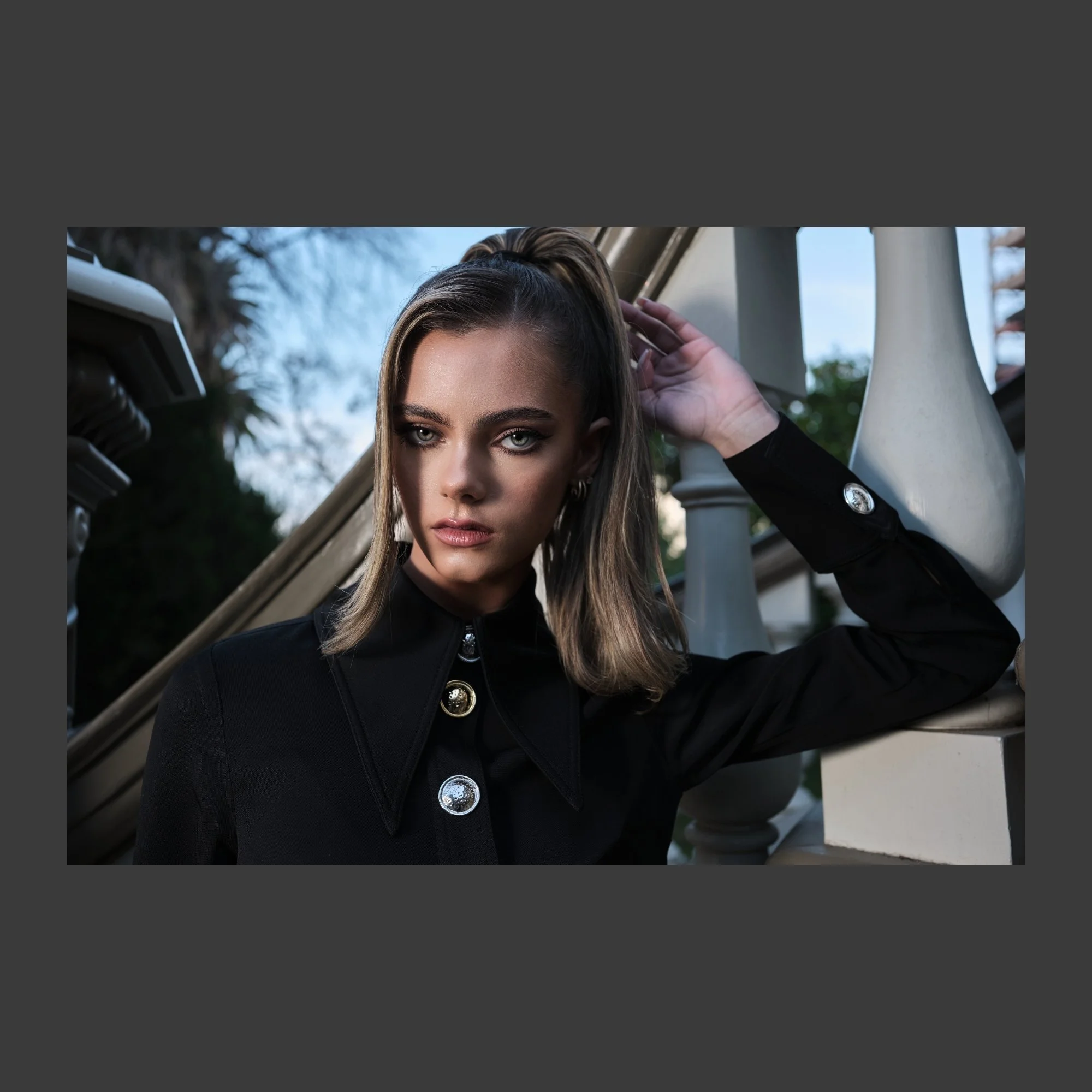 Young woman with shoulder-length hair, wearing a black coat with metallic buttons, posing outside near an ornate staircase.