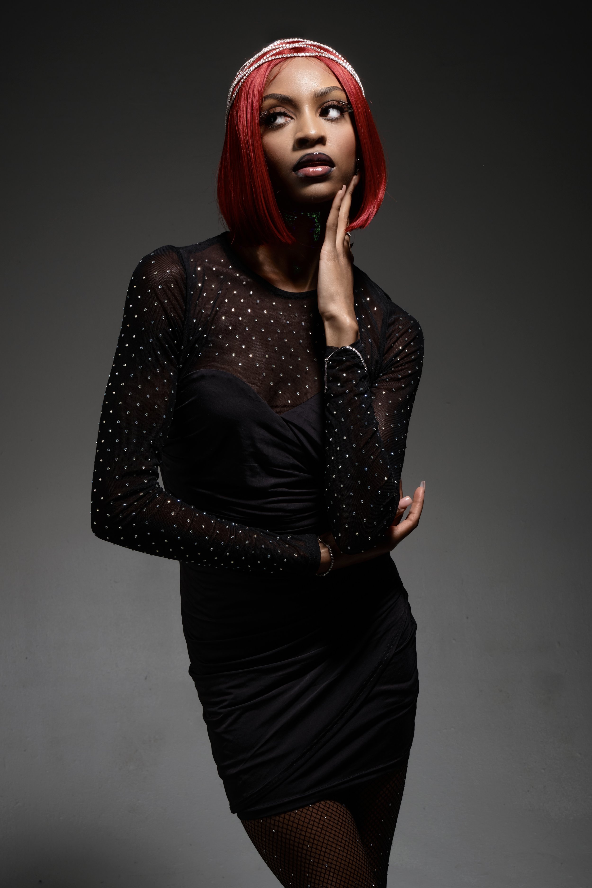 A woman with red hair wearing a black, sheer, long-sleeved top with rhinestones, a black dress, fishnet tights, and a silver headband with rhinestones, posing against a gray background.
