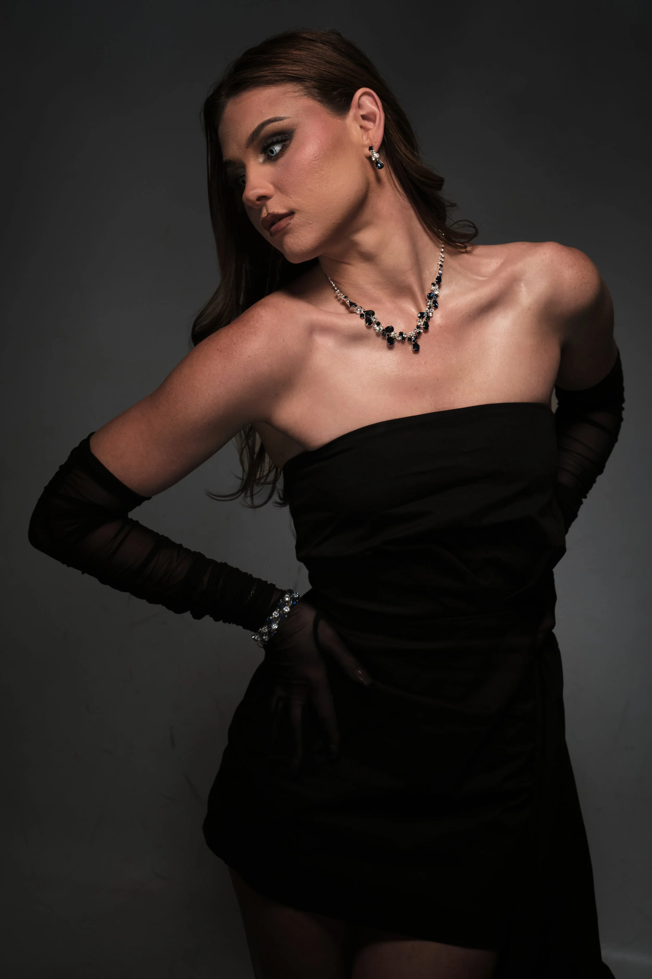 A woman wearing a strapless black dress, long black gloves, and jewelry posed against a dark background.