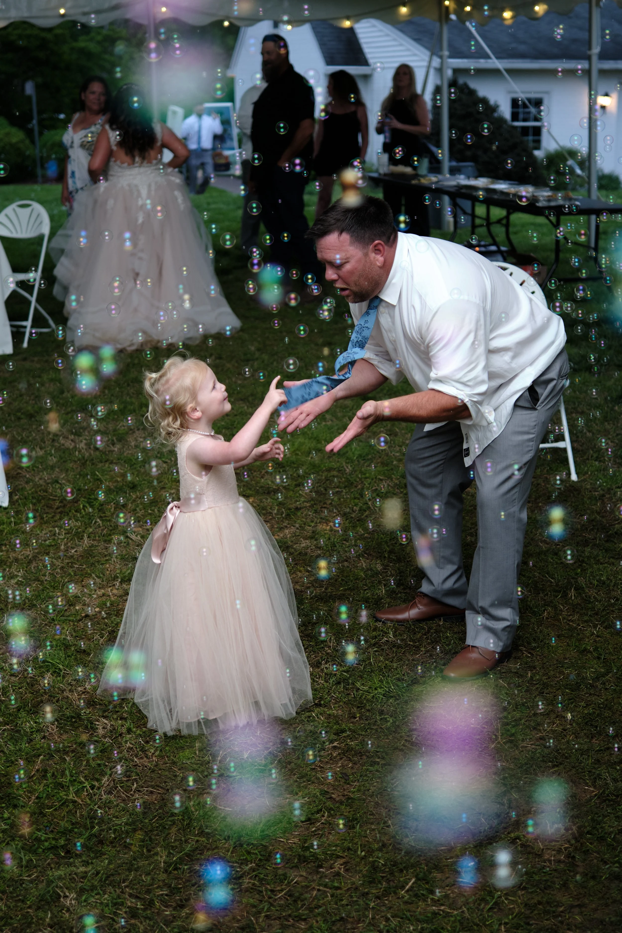 A man and a young girl dancing together at an outdoor wedding reception during the evening, surrounded by bubbles and other guests in the background.