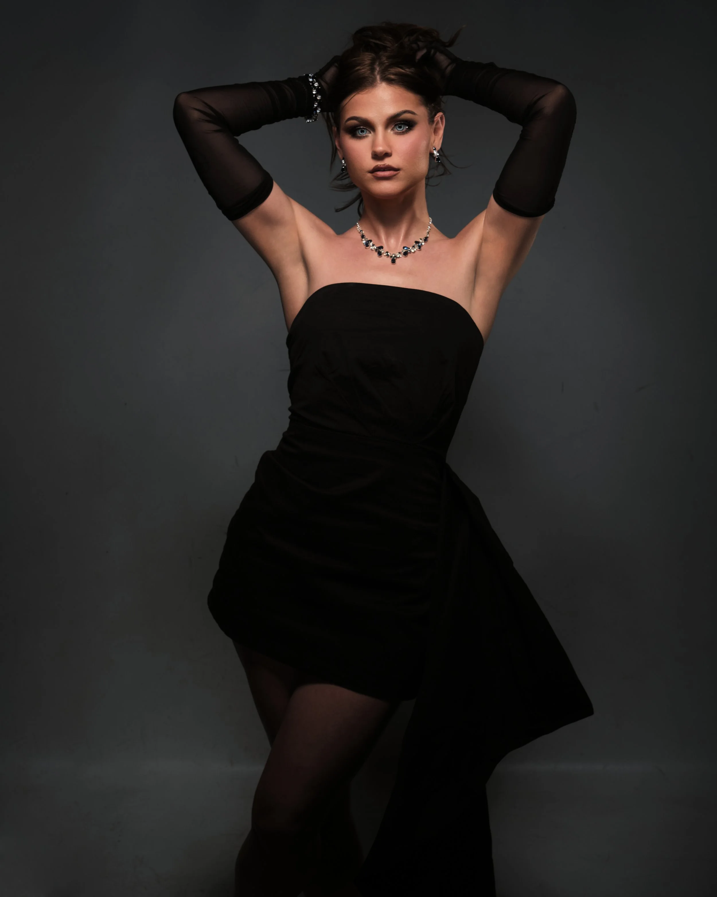 A woman wearing a strapless black dress and black sheer gloves, posing with her hands behind her head, against a dark background.