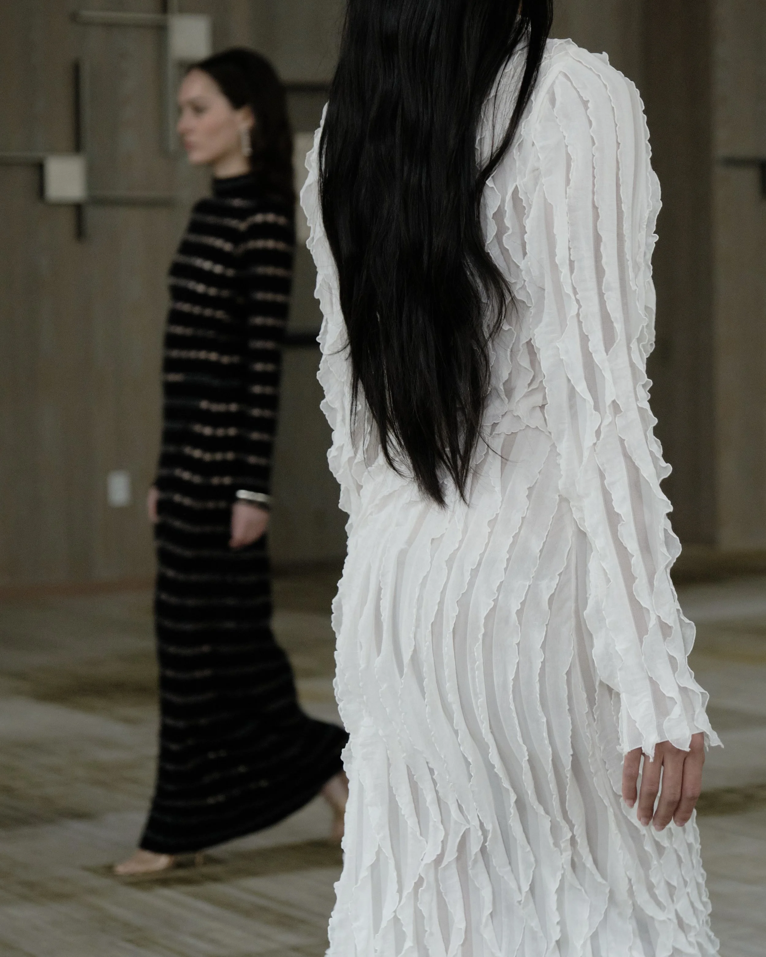 A group of models walking down a runway during a fashion show, wearing various designer outfits in a large indoor venue with wooden paneling.