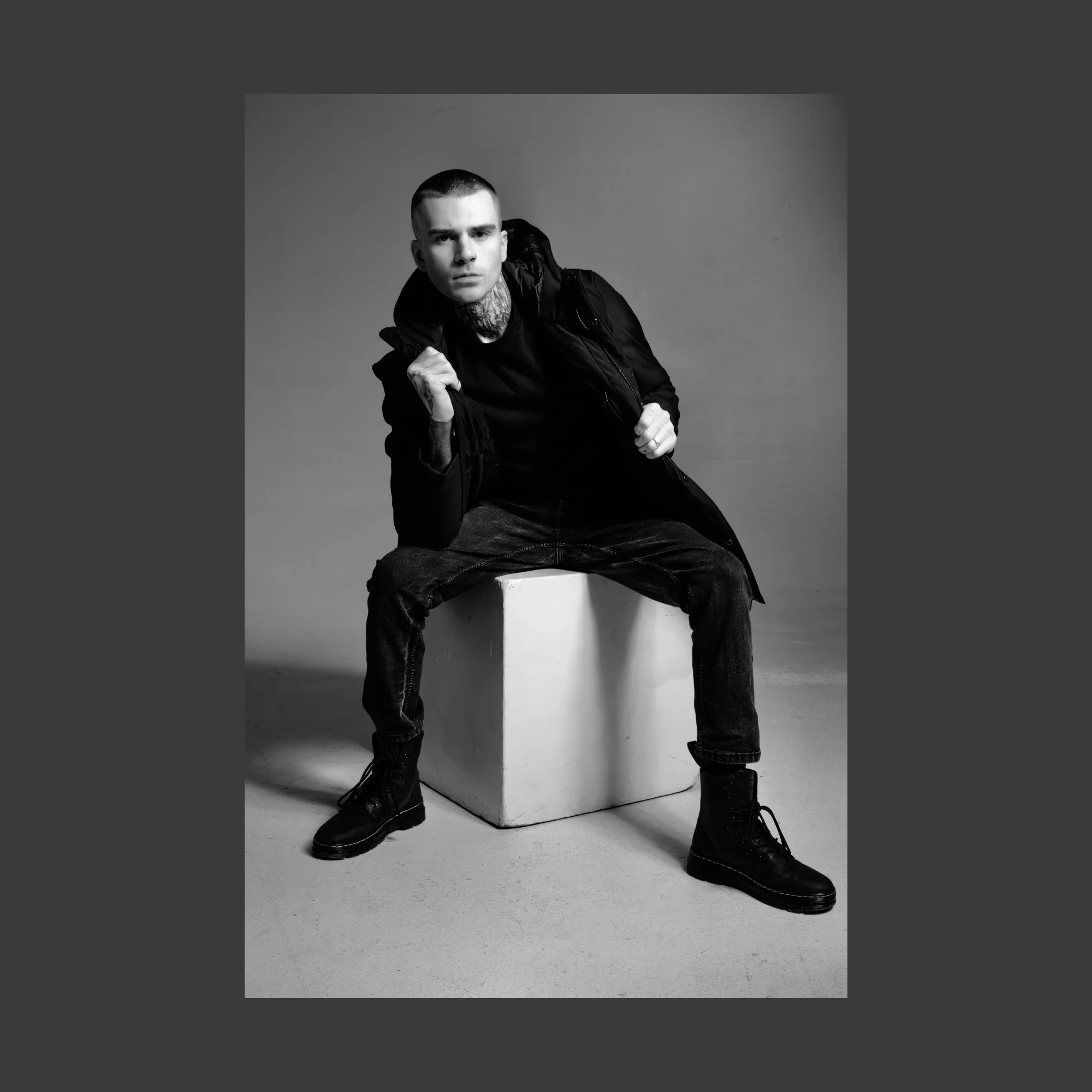 Black and white photo of a young man sitting on a cube, wearing dark clothing, boots, and a jacket, posing with fists clenched and a serious expression.