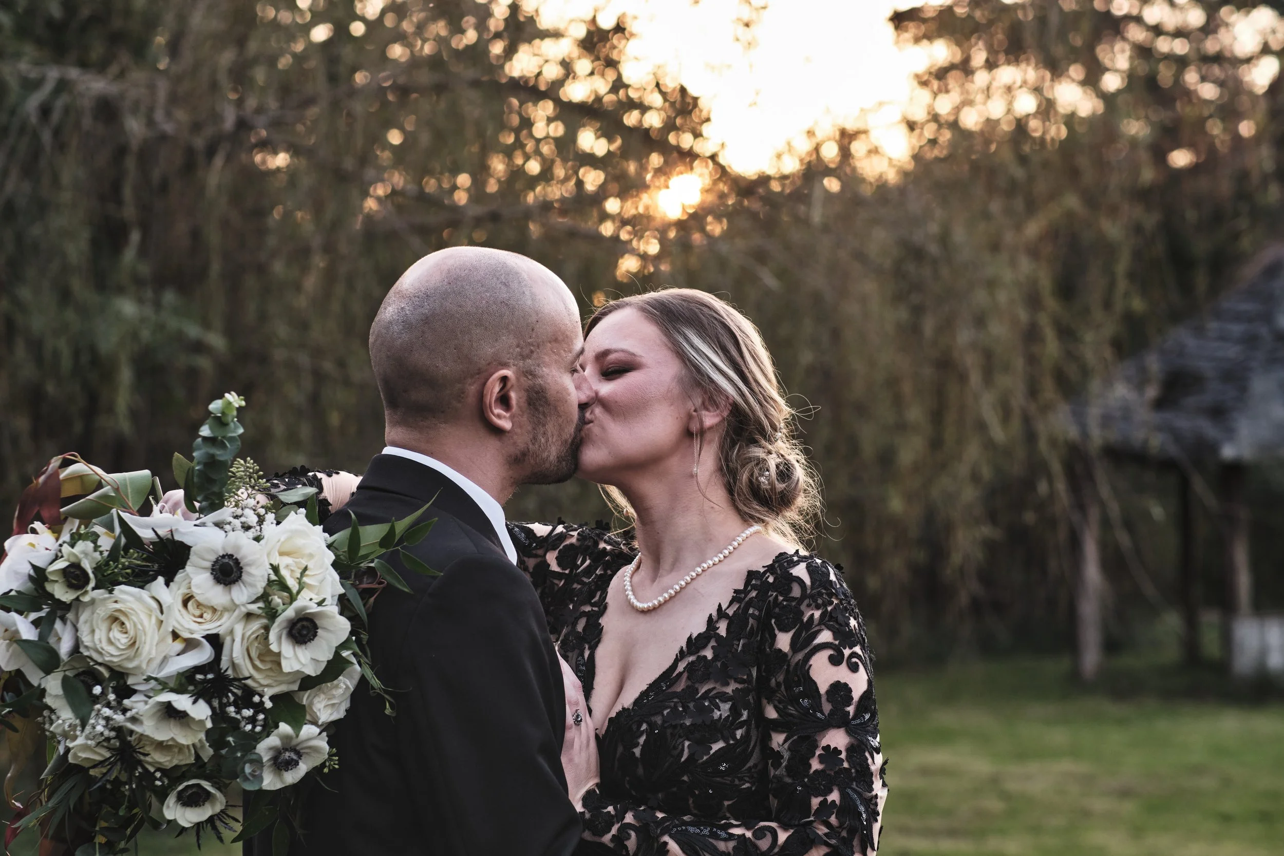 A couple in wedding attire sharing a kiss outdoors at sunset, with trees and a grassy area in the background.