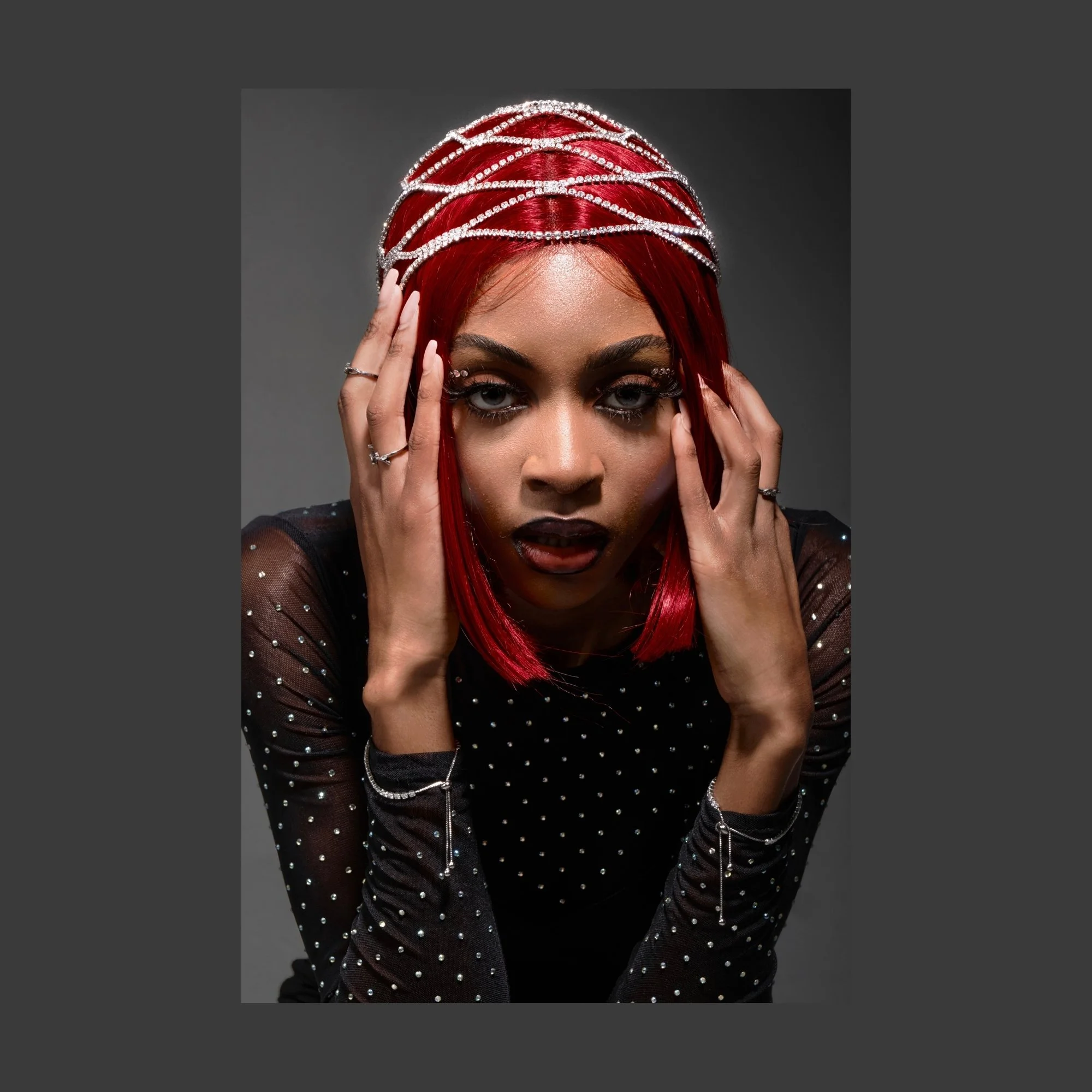A woman with bright red hair and striking makeup, wearing a black mesh top with rhinestone embellishments, holding her head with both hands, against a gray background.