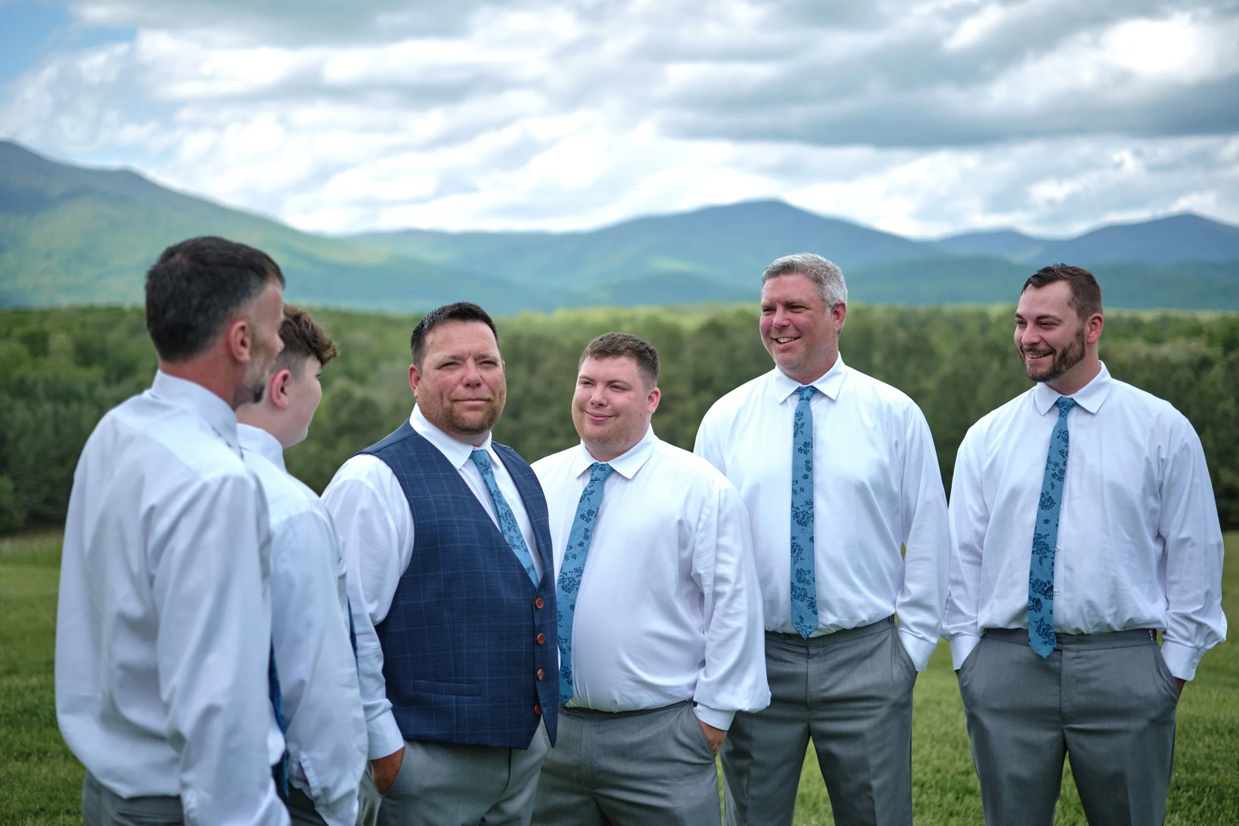 Group of five men dressed in white shirts, gray pants, and blue ties, standing outdoors in a green field with mountains and cloudy sky in the background, engaging in conversation and smiling.