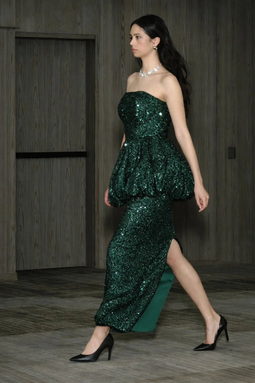 Woman in a strapless, emerald green, glittery gown with a high slit, black high heels, silver jewelry, and dark hair walking in a room with wooden walls.