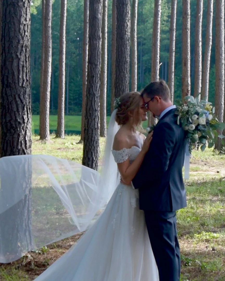 A bride and groom standing close together in a forest, touching foreheads, with the bride holding a bouquet of white roses and greenery.