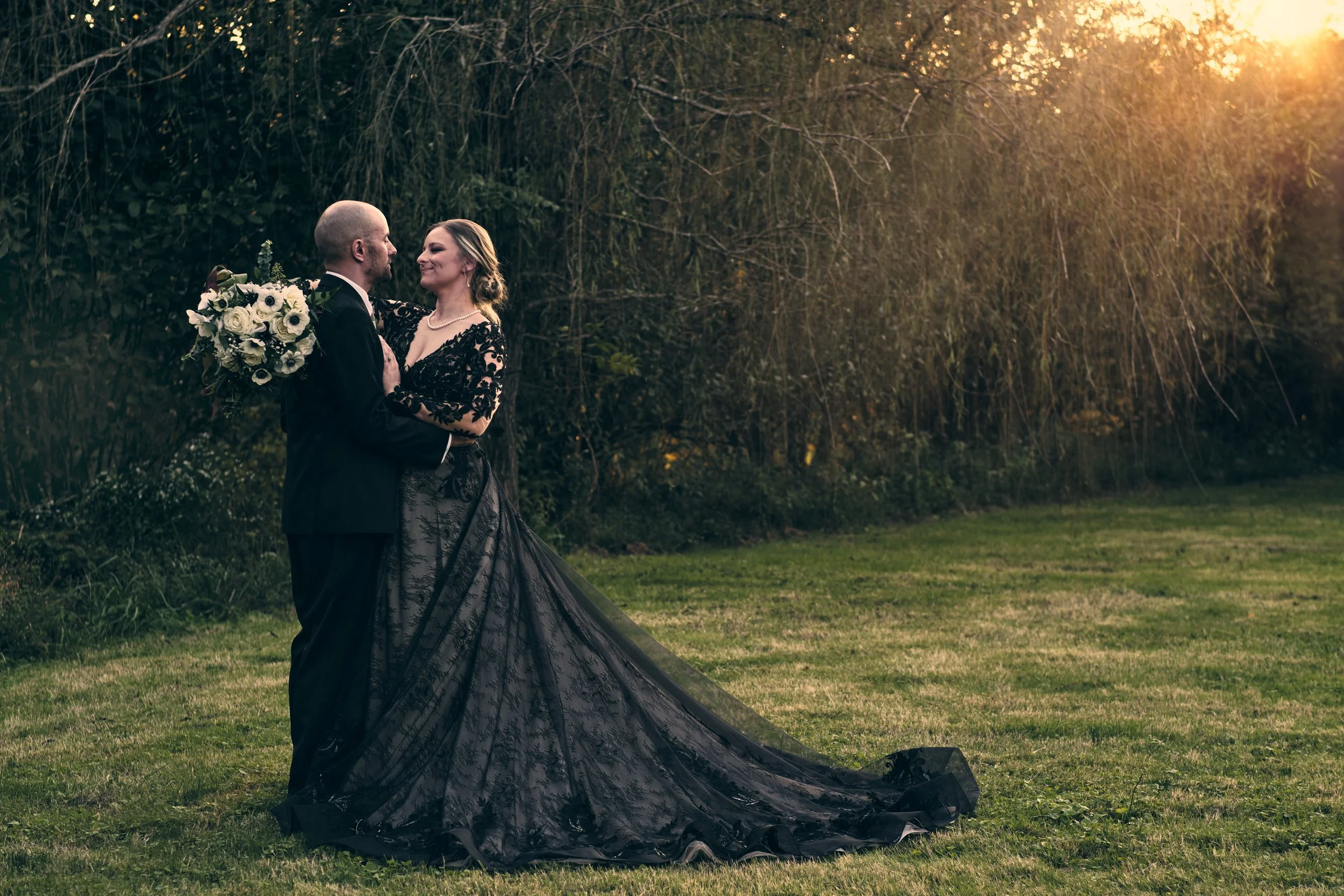 A bride and groom dance outdoors during sunset, surrounded by trees, with the bride holding a bouquet of white flowers and wearing a black lace gown.