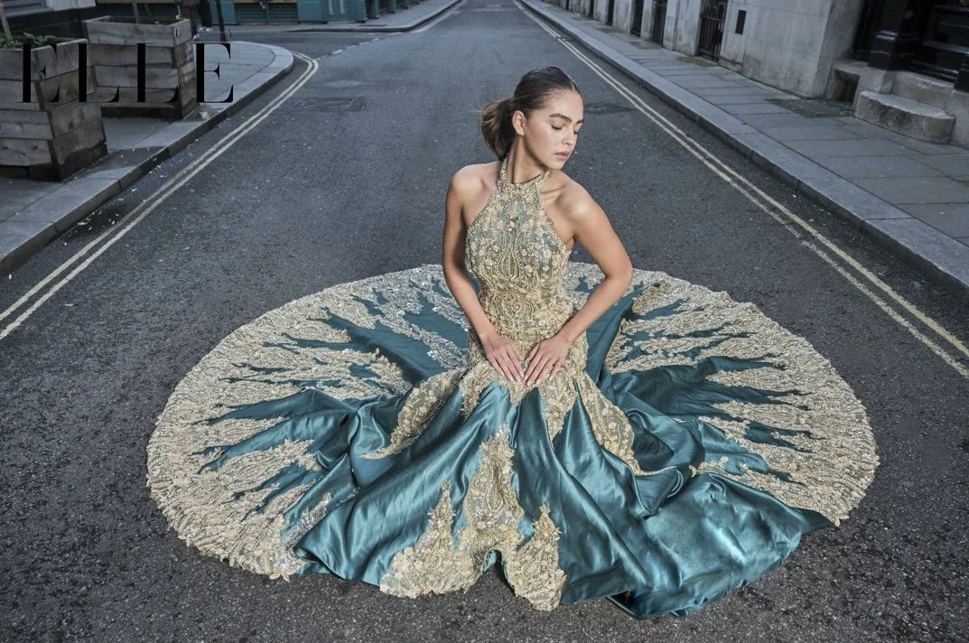 A woman sitting on an empty city street wearing an ornate teal and gold gown that fans out around her like a butterfly or flower.