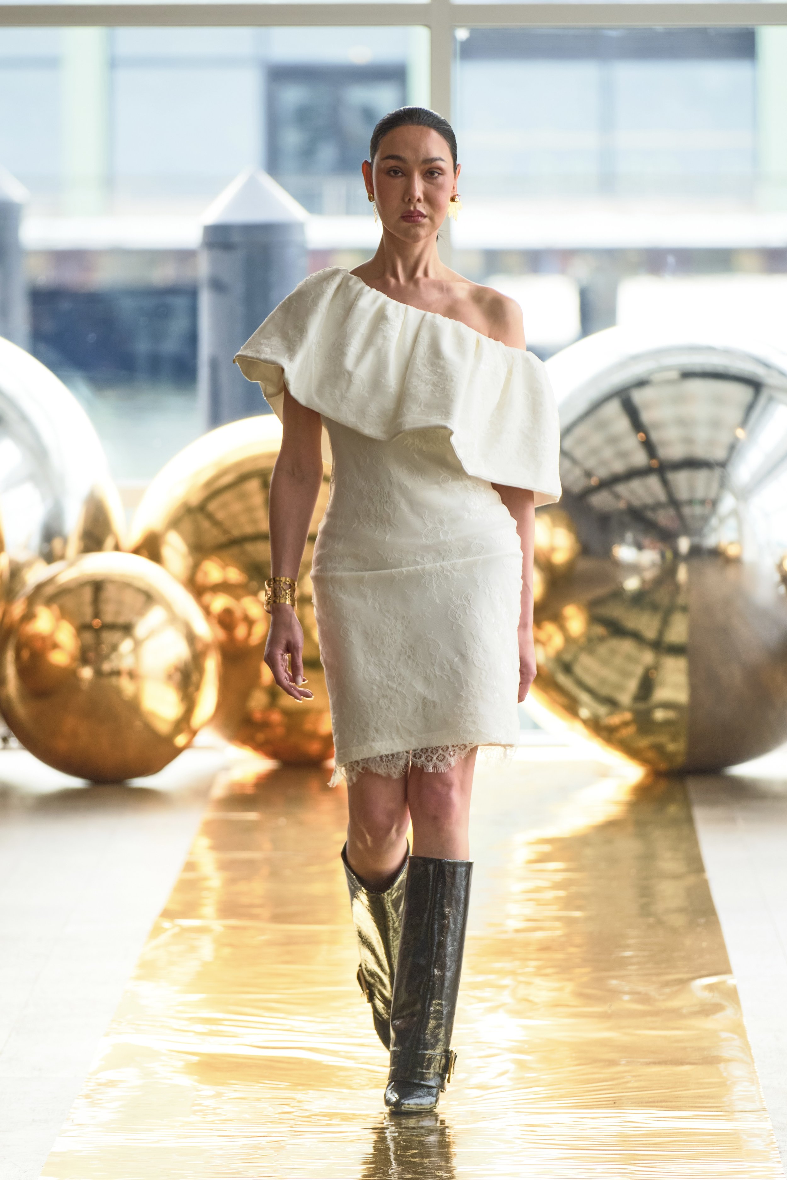 Woman walking on a gold runway with large gold balls in the background, wearing a cream-colored dress with one shoulder ruffle and gold knee-high boots.