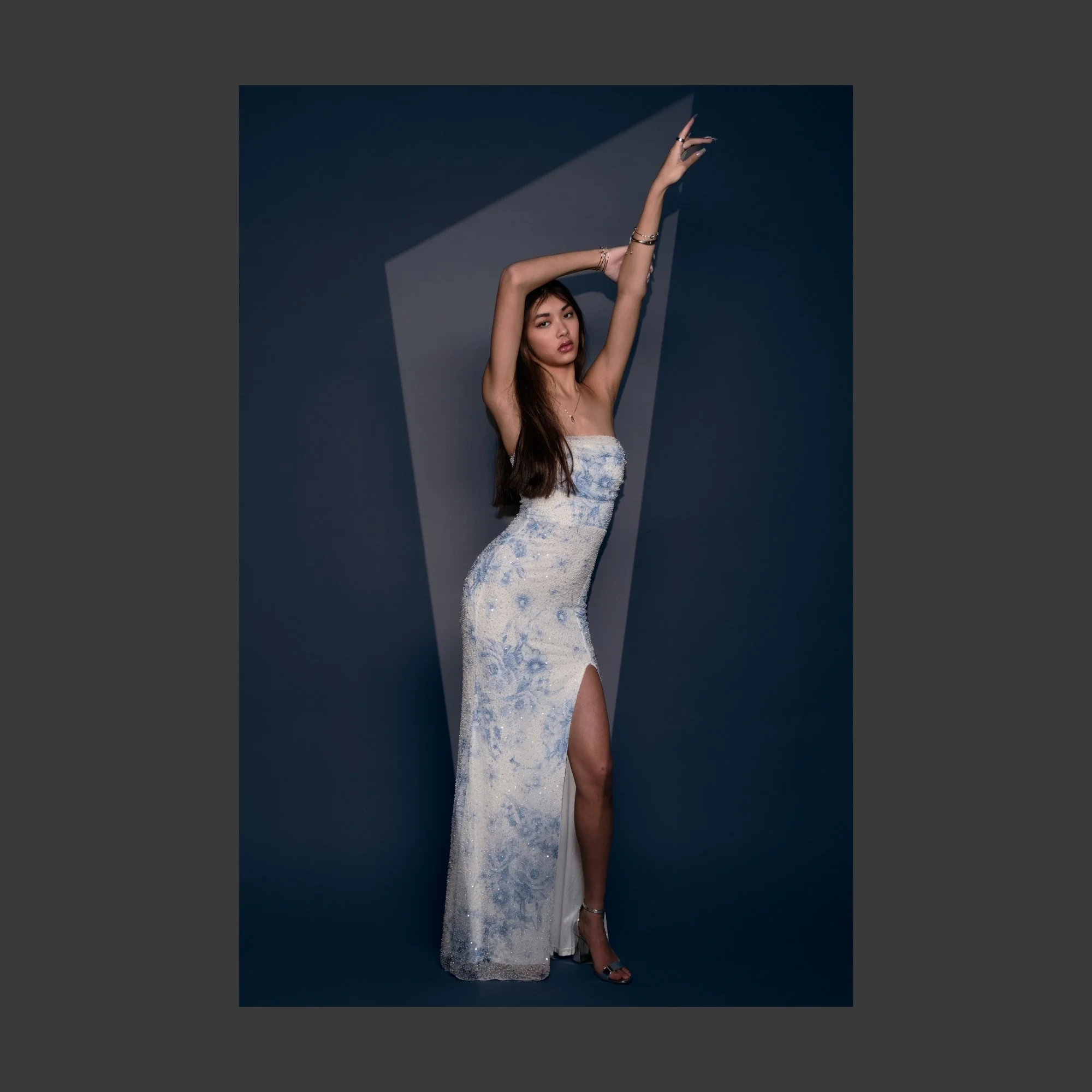 A young woman wearing a strapless, high-slit, floor-length white dress with blue floral details, standing against a blue background with a geometric cutout, striking a pose with one arm raised above her head and the other bent, showcasing her jewelry