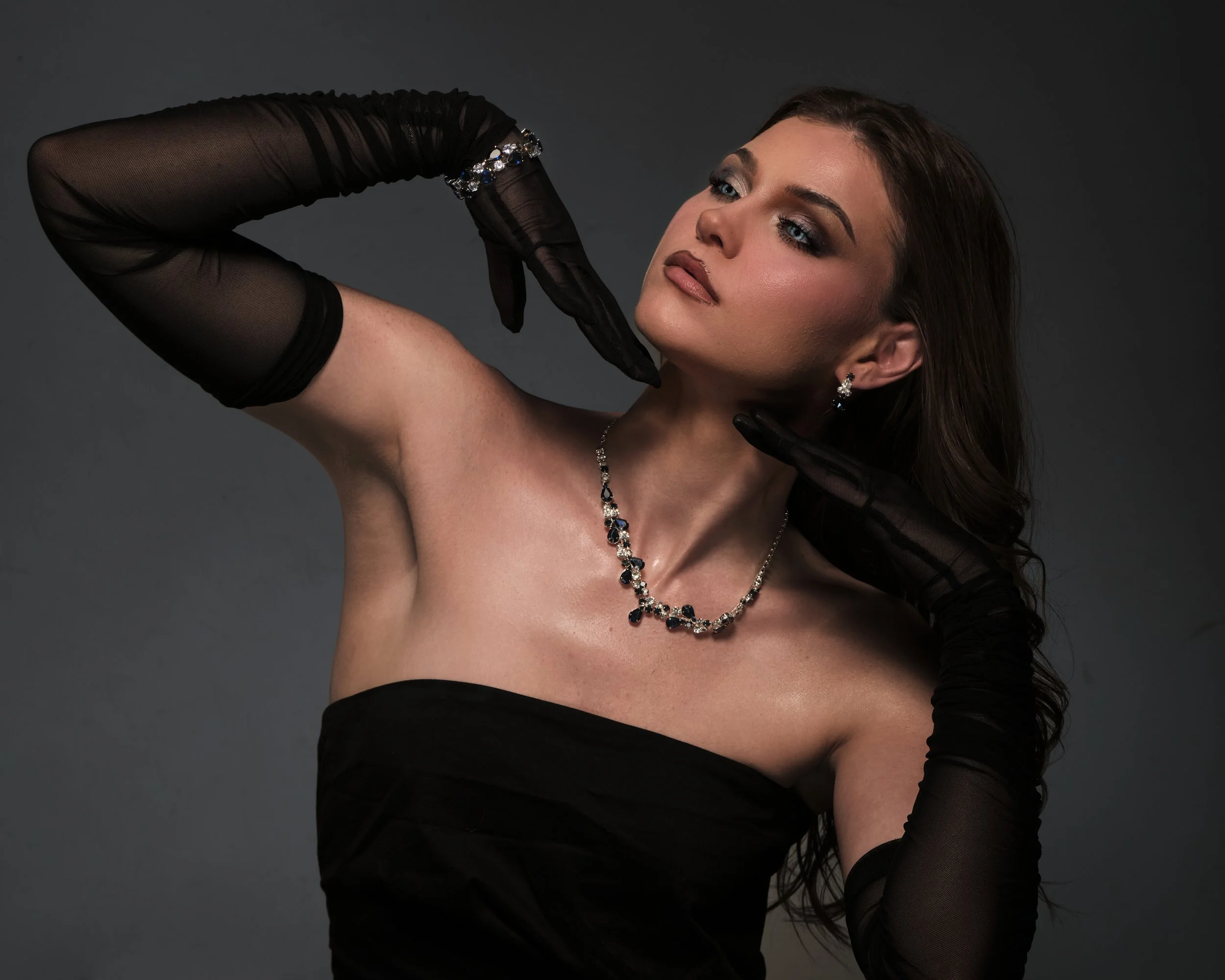 A woman wearing elegant jewelry and black gloves poses against a dark background.
