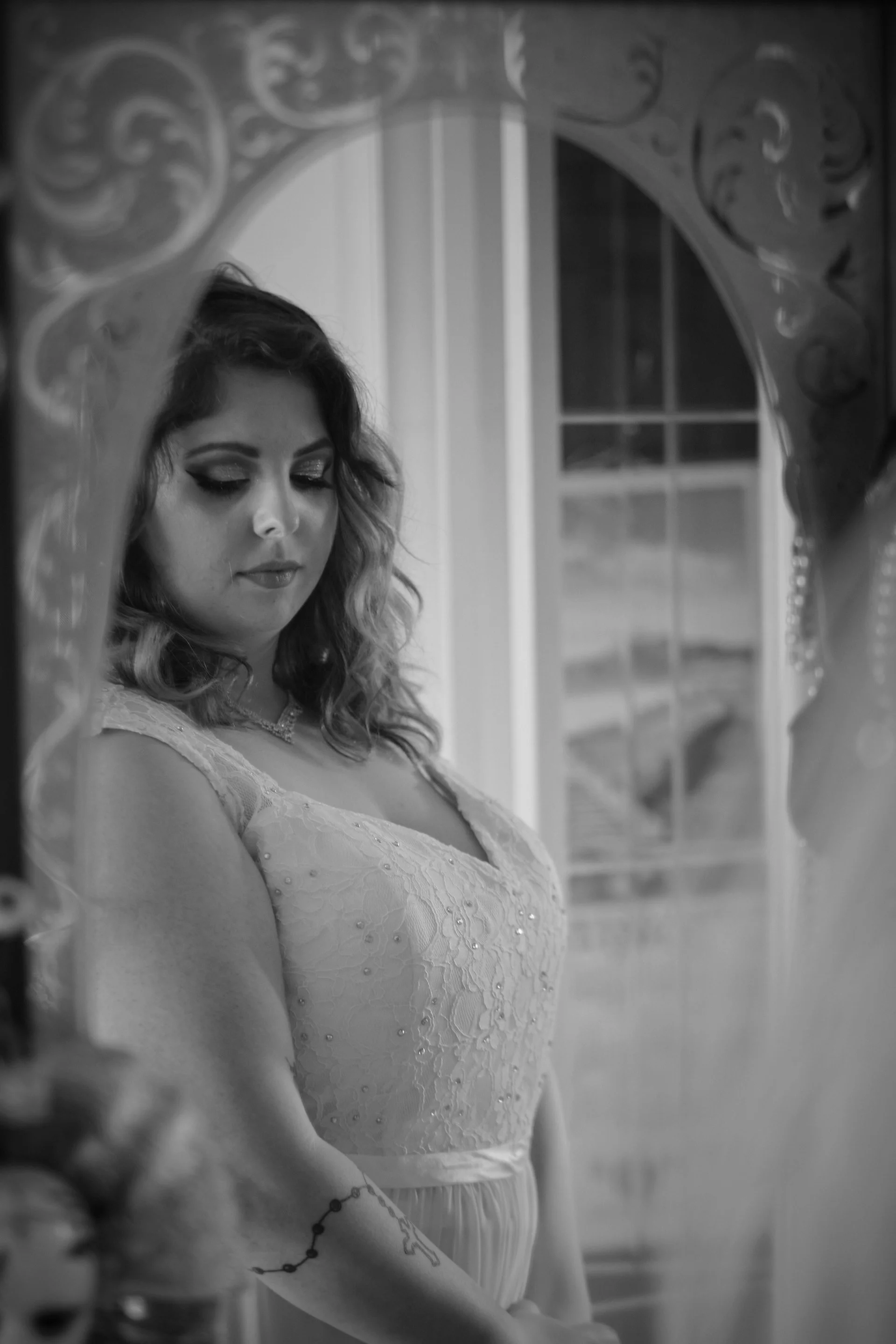 A woman with styled hair and makeup, wearing a lace dress with embellishments, looking down, seen through a decorative mirror in black and white.