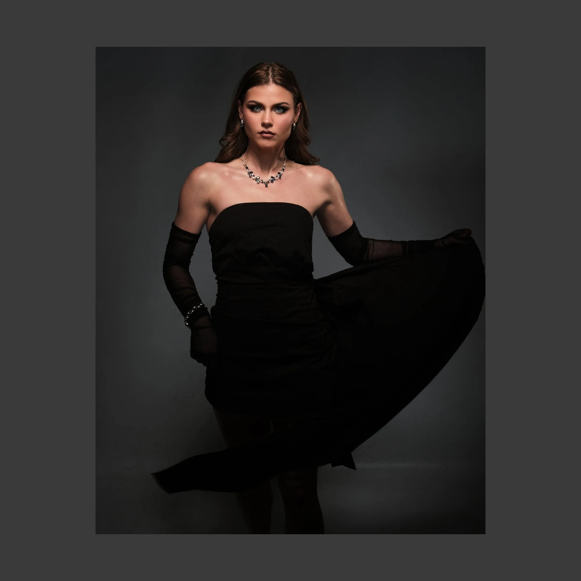 A woman with brown hair and blue eyes in a strapless black evening gown, wearing black gloves, a pearl necklace, and earrings, standing against a dark background.