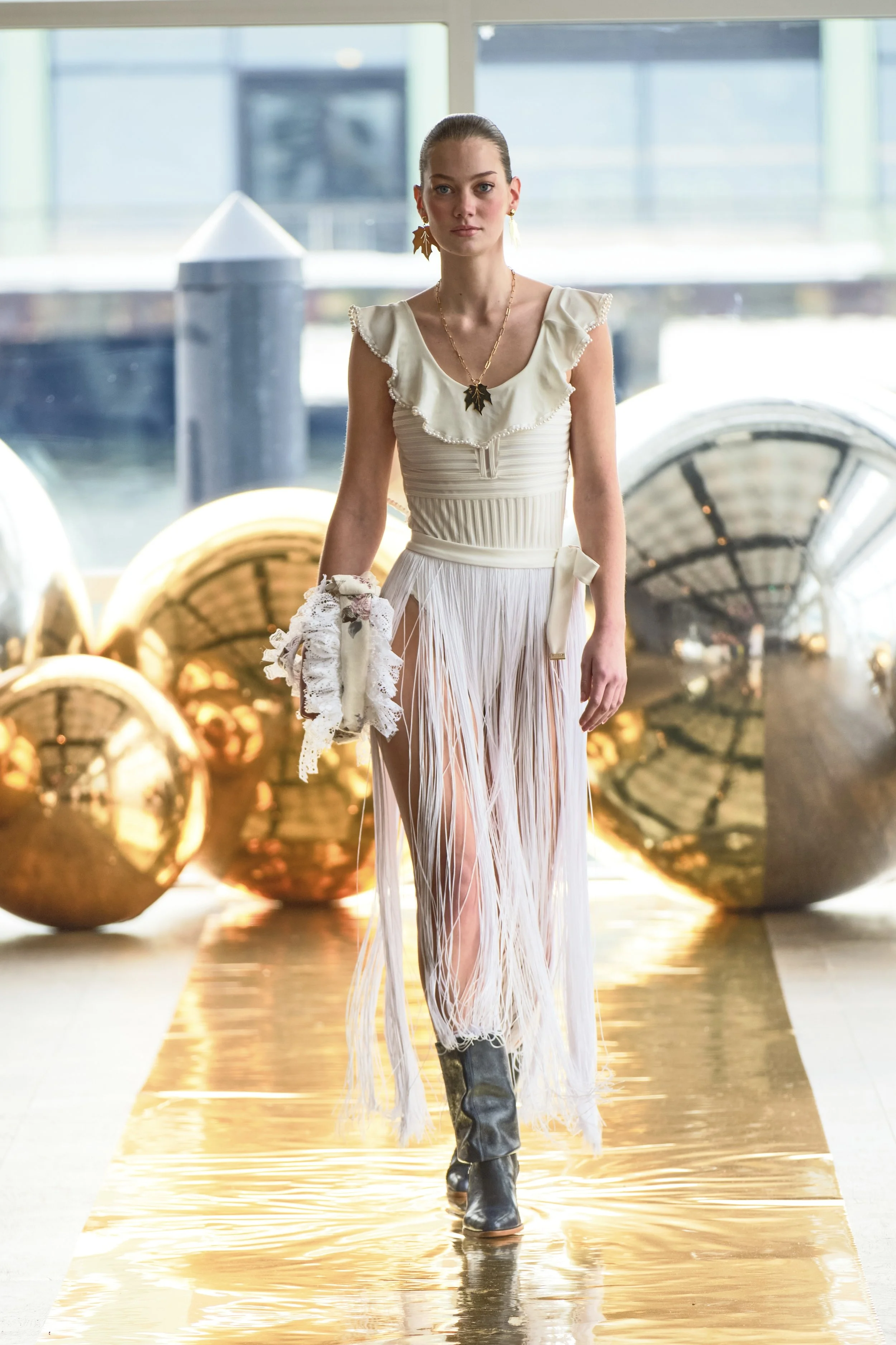 A female model walking along a golden runway in a fashion show, wearing a white ruffled top and a long, fringed skirt with high slit, black boots, and accessories including a leaf-shaped necklace and matching earrings and gloves.