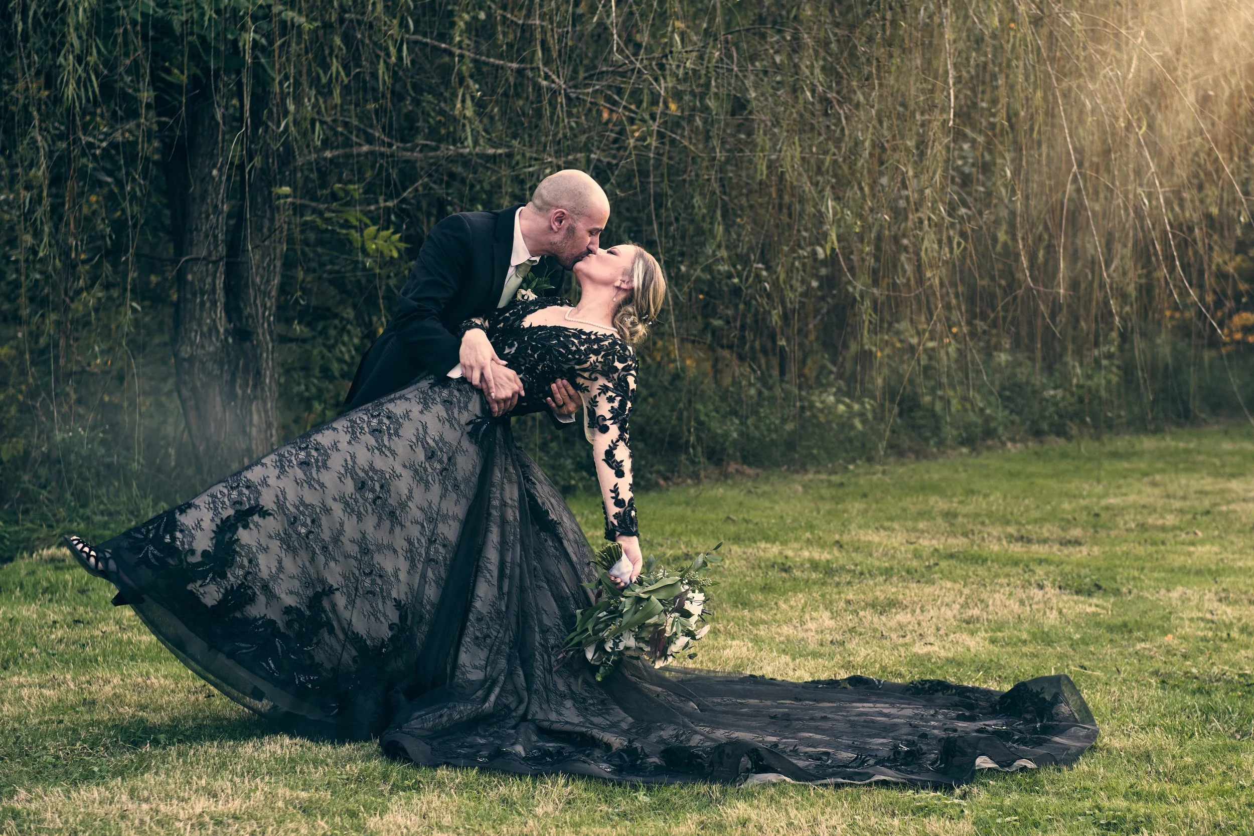 A newlywed couple sharing a kiss outdoors, with the groom dipping the bride, who is holding a bouquet of flowers, on a grassy area with trees in the background.