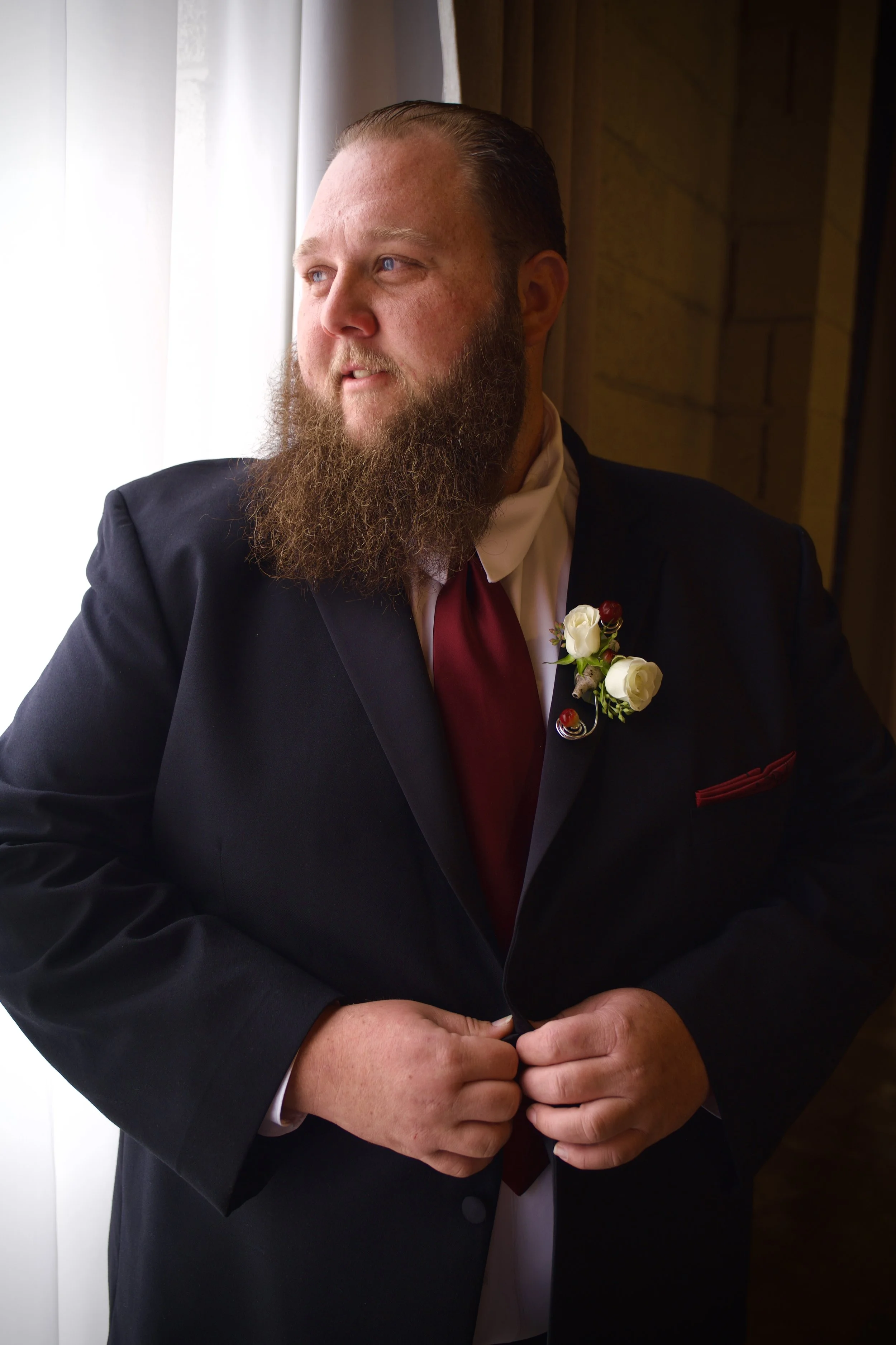 Man with a long beard in a black tuxedo standing near a window with light coming in.